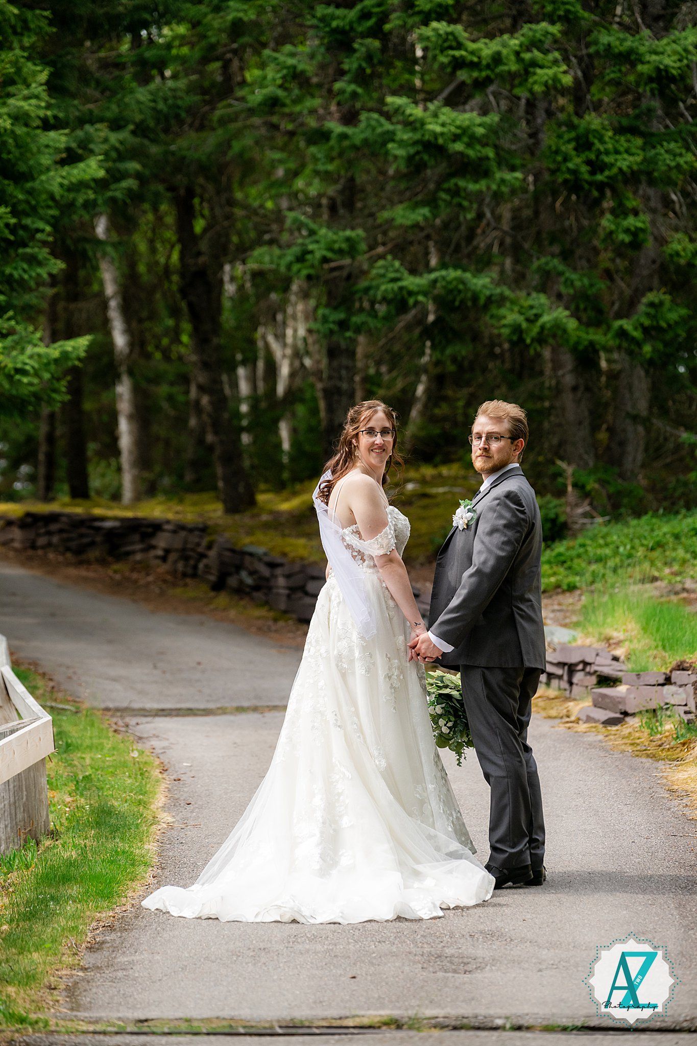 A newlywed couple poses on a scenic path surrounded by lush green trees and a stone wall on their wedding day.