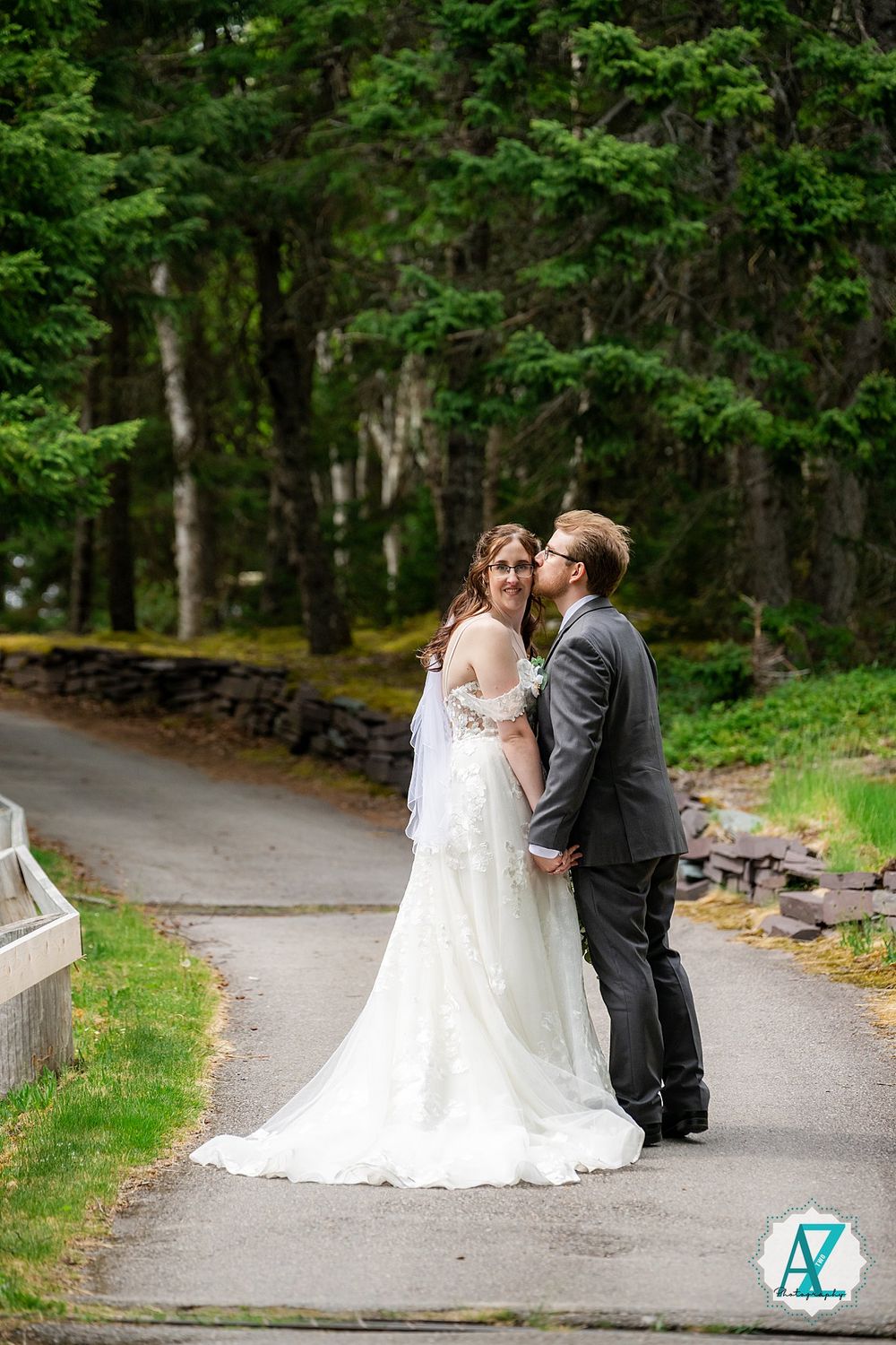 A couple shares a romantic moment on a tree-lined path in formal wedding attire surrounded by lush greenery.