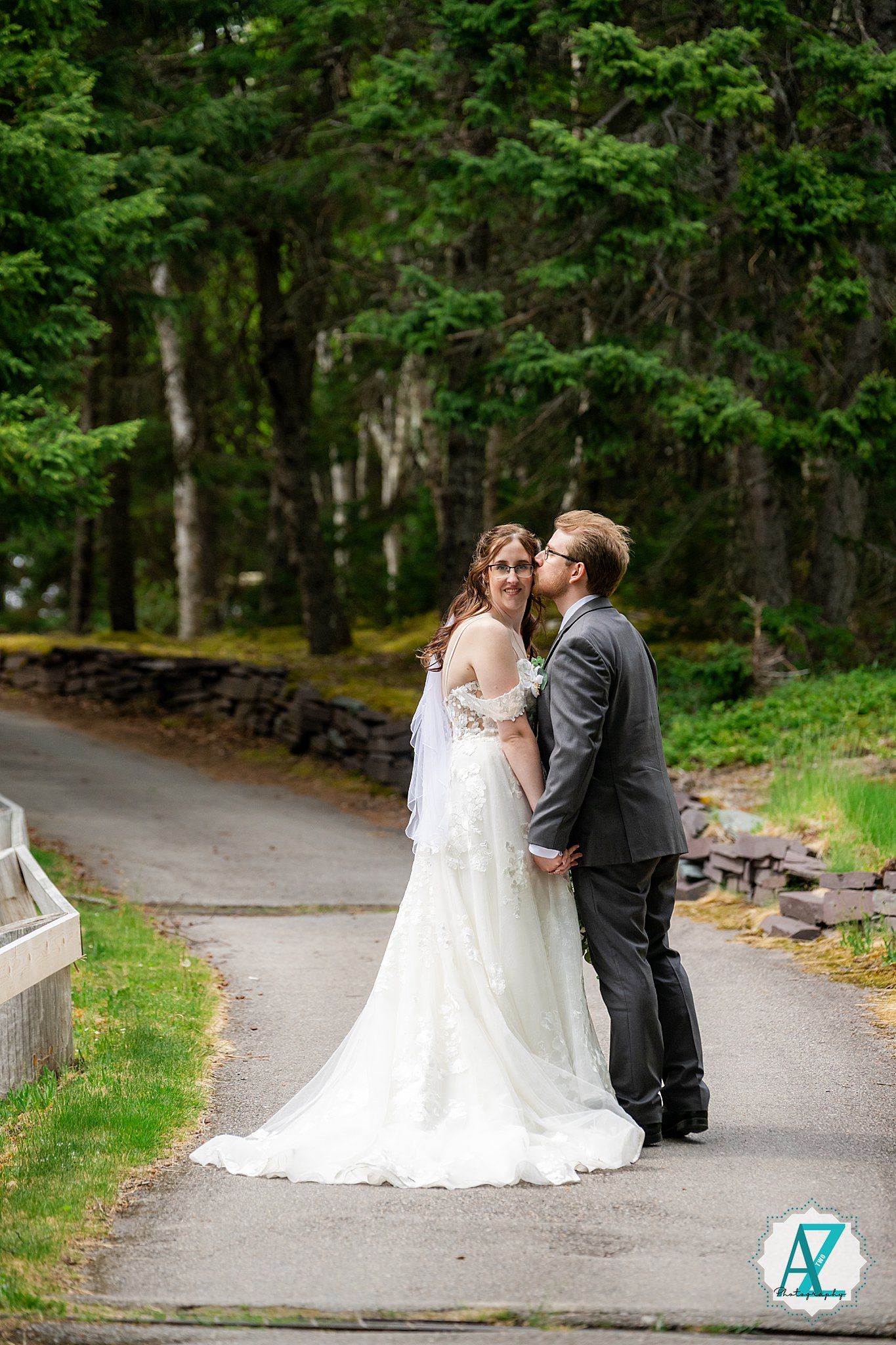 A couple shares a romantic moment on a tree-lined path in formal wedding attire surrounded by lush greenery.