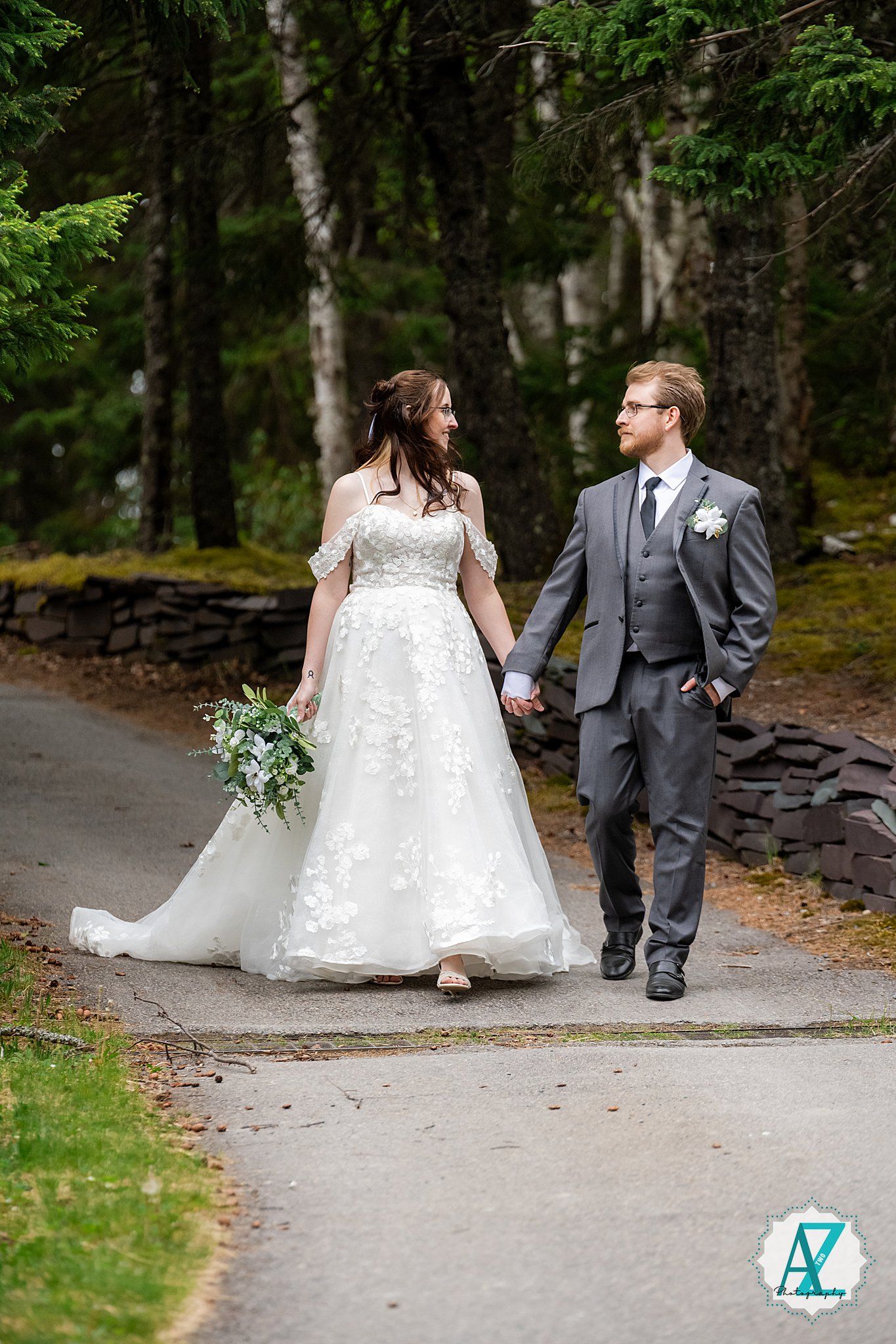 A couple in wedding attire walks together on a stone-lined path through a lush wooded area.