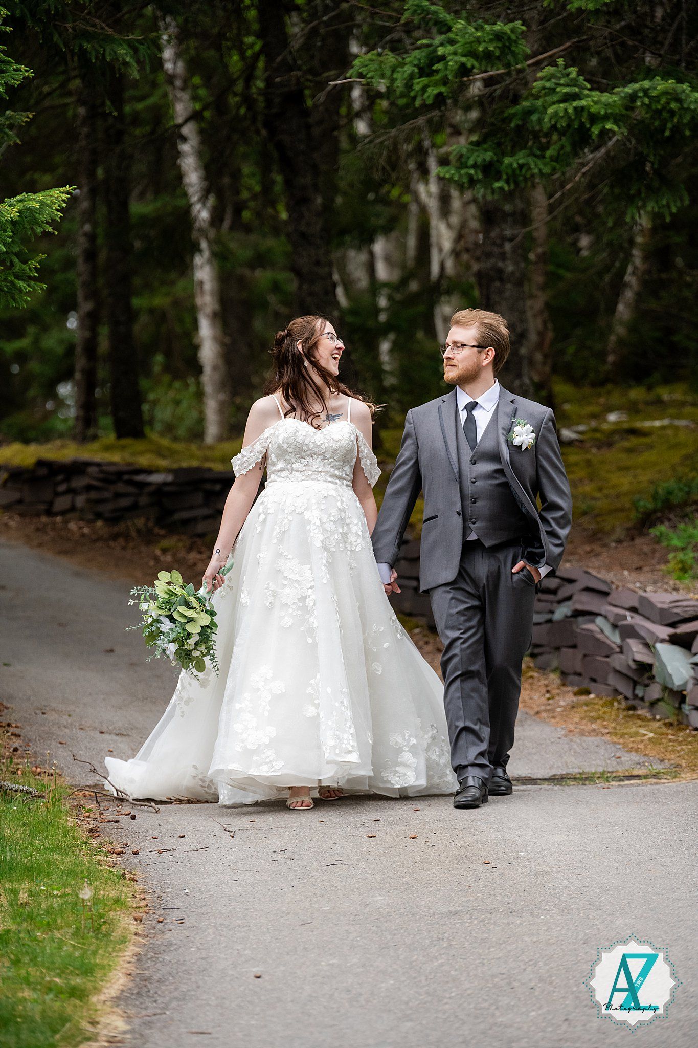 A newlywed couple walks together on a wooded path, with the bride in a lace gown and groom in a gray suit.