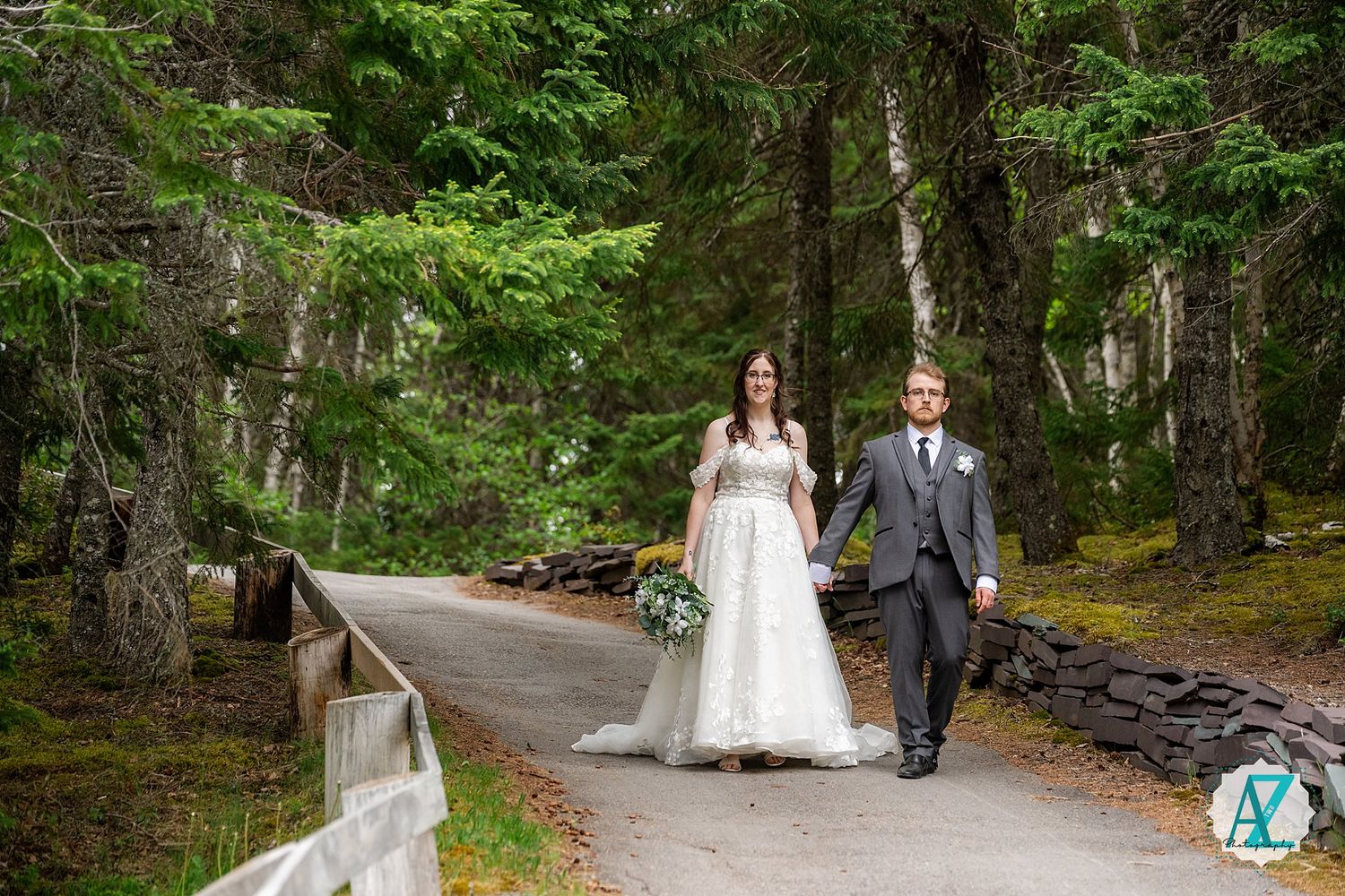 A couple walks hand in hand down a forest path dressed in wedding attire surrounded by tall evergreen trees.