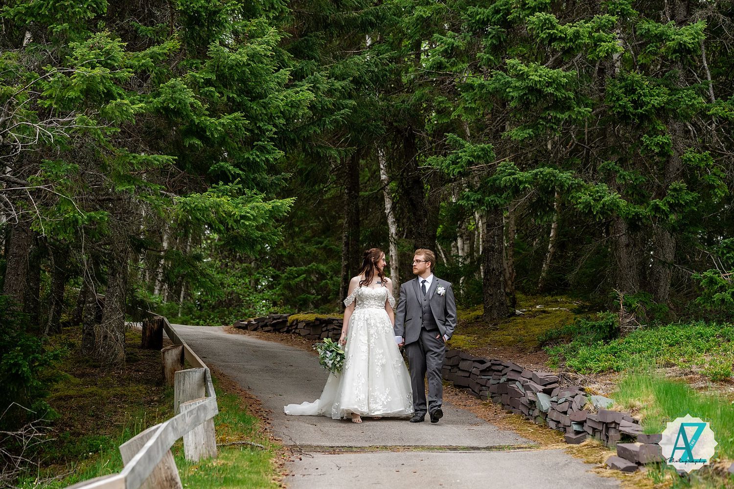 A couple walks along a scenic wooded path lined with stone walls and lush greenery on a bright summer day.