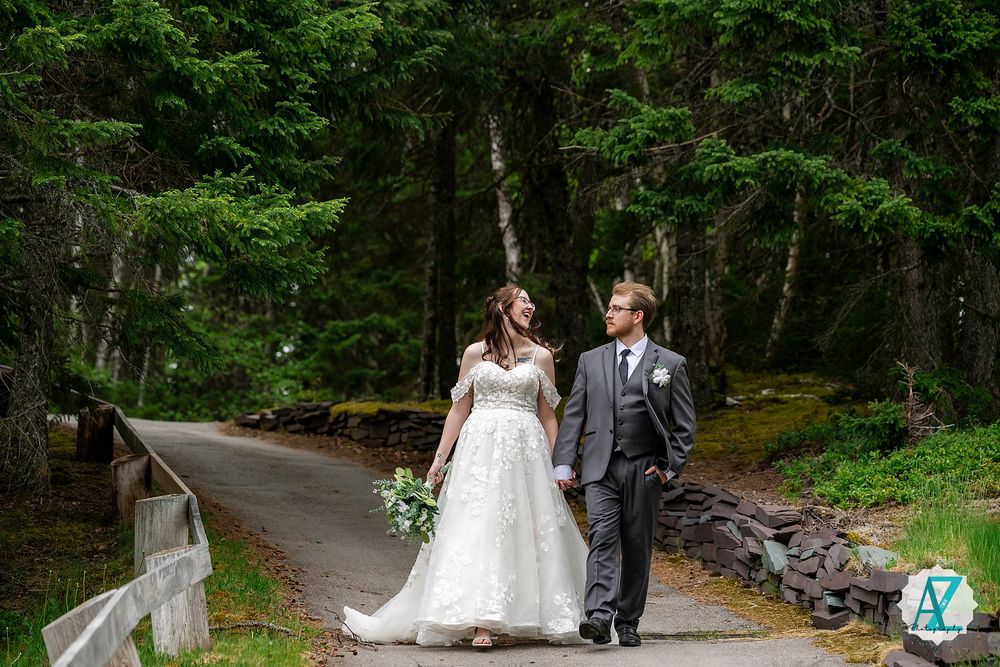 A newlywed couple walks hand in hand along a scenic forest path surrounded by lush green trees.