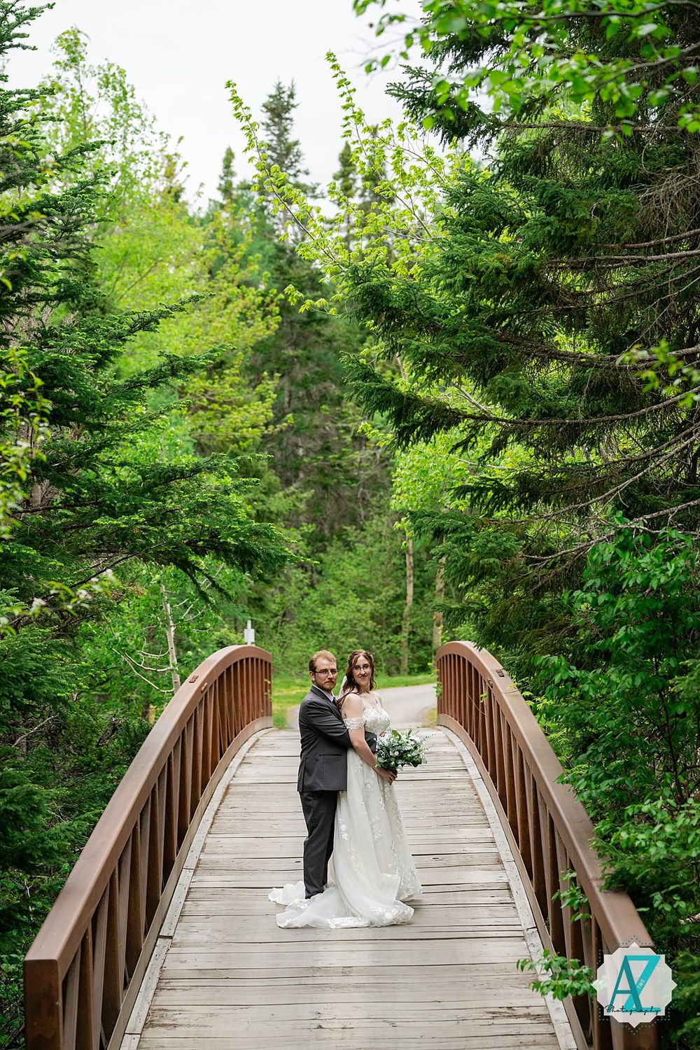 A couple stands on a wooden footbridge surrounded by lush evergreen forest on their wedding day.