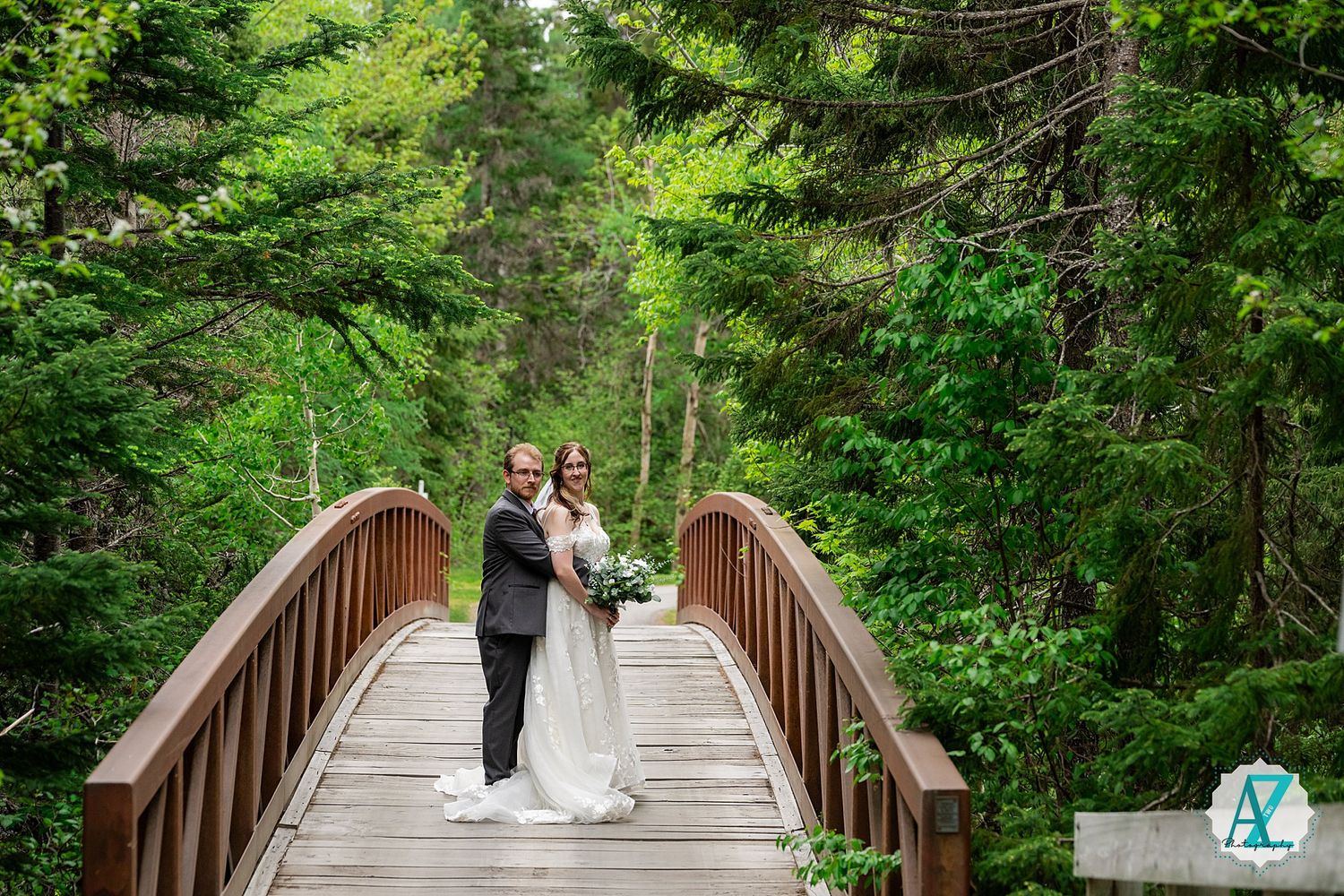 A romantic wedding photo captured on a wooden bridge with towering evergreens in the background.