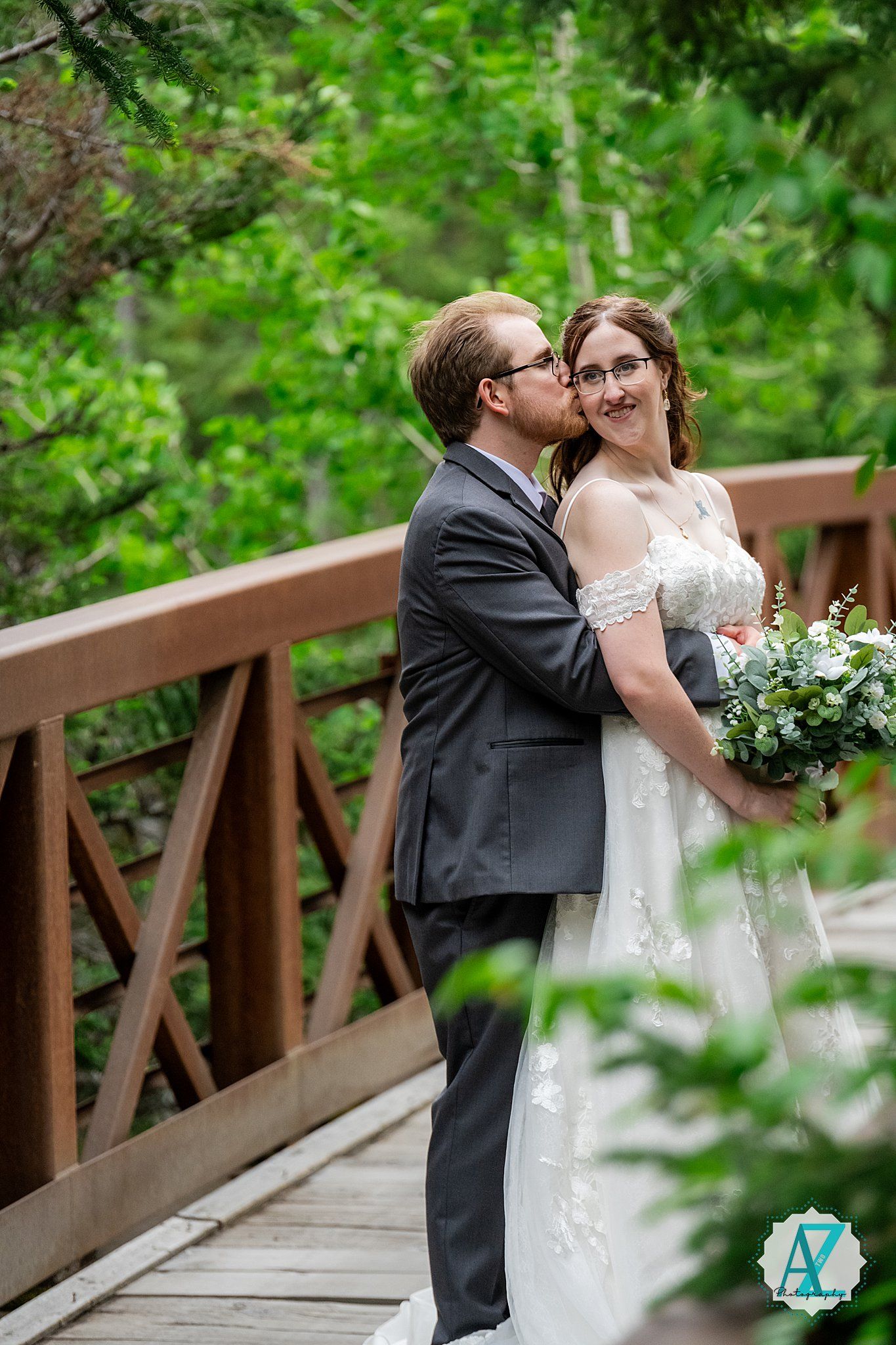 Newlywed couple shares intimate moment on wooden bridge surrounded by lush green foliage with white floral bouquet.