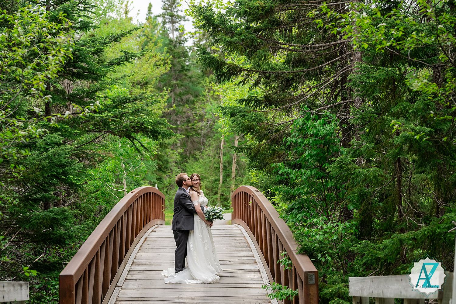 Wooden footbridge crosses through lush green forest with couple standing in peaceful natural setting.