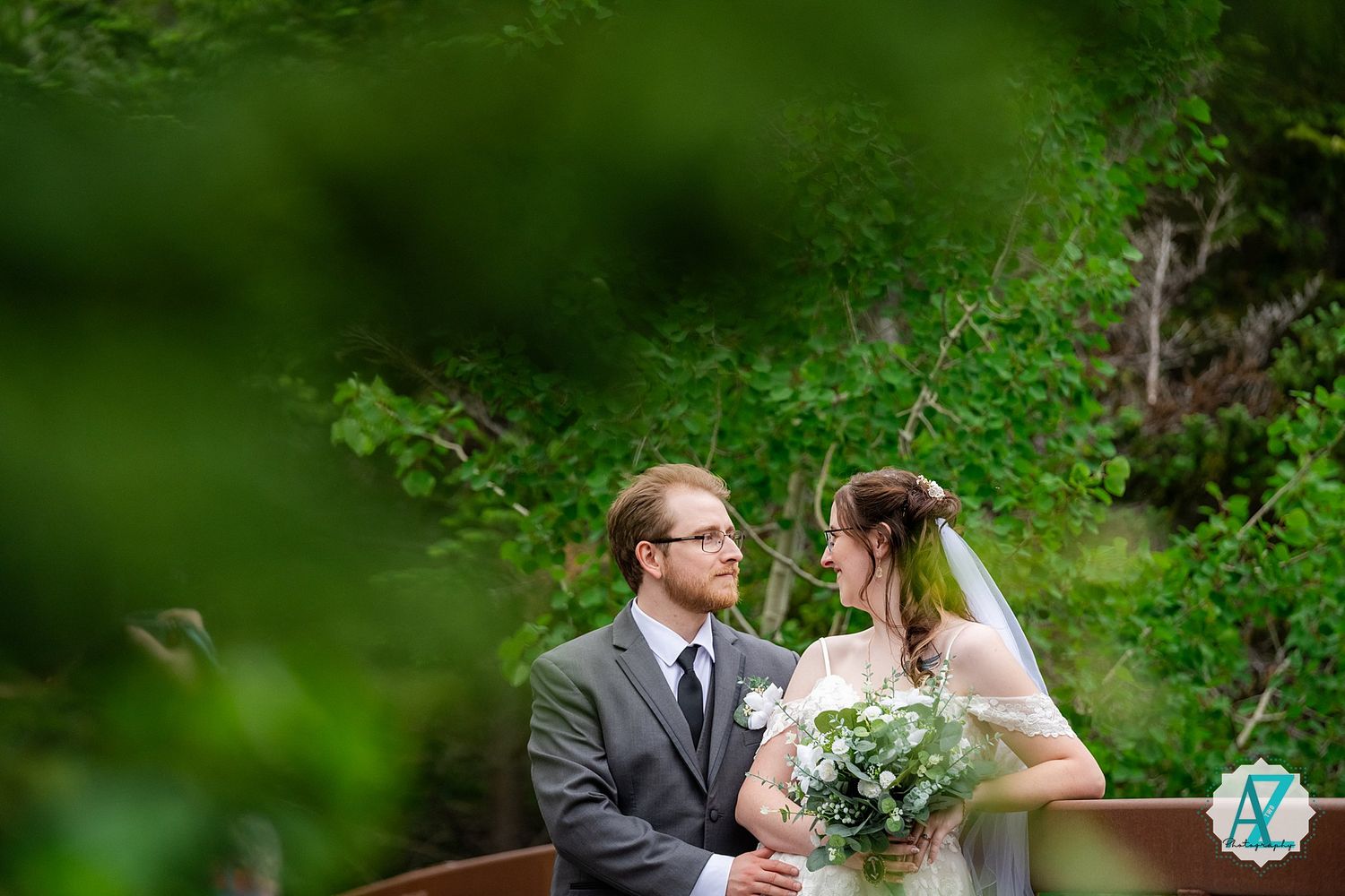 A couple sits together on a wooden bench surrounded by lush green foliage during their outdoor wedding ceremony.