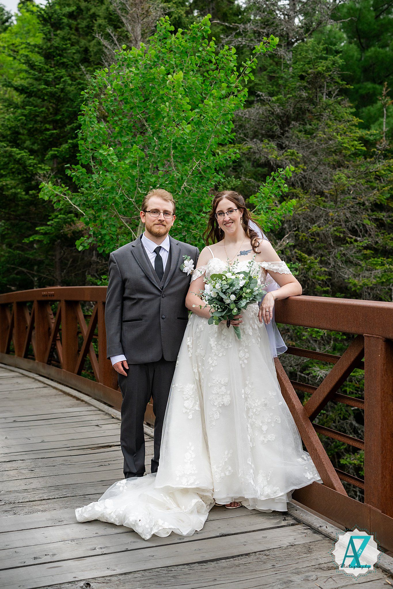 A wedding couple stands on a wooden bridge surrounded by lush green forest scenery.