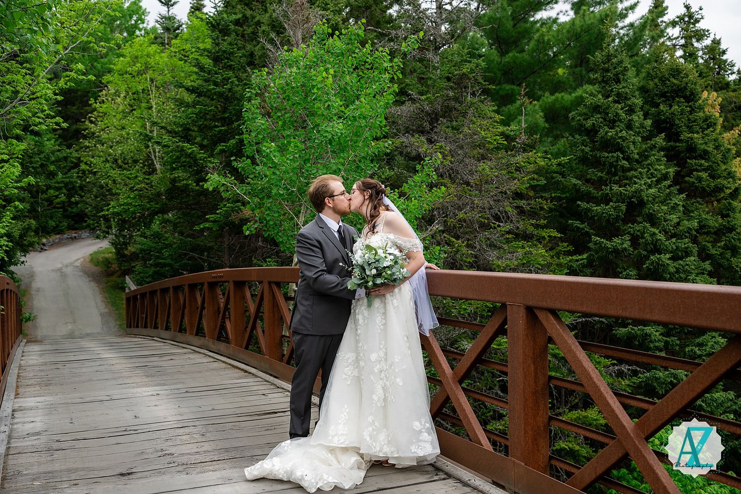 A couple in wedding attire stands on a rustic wooden bridge surrounded by lush evergreen forest.