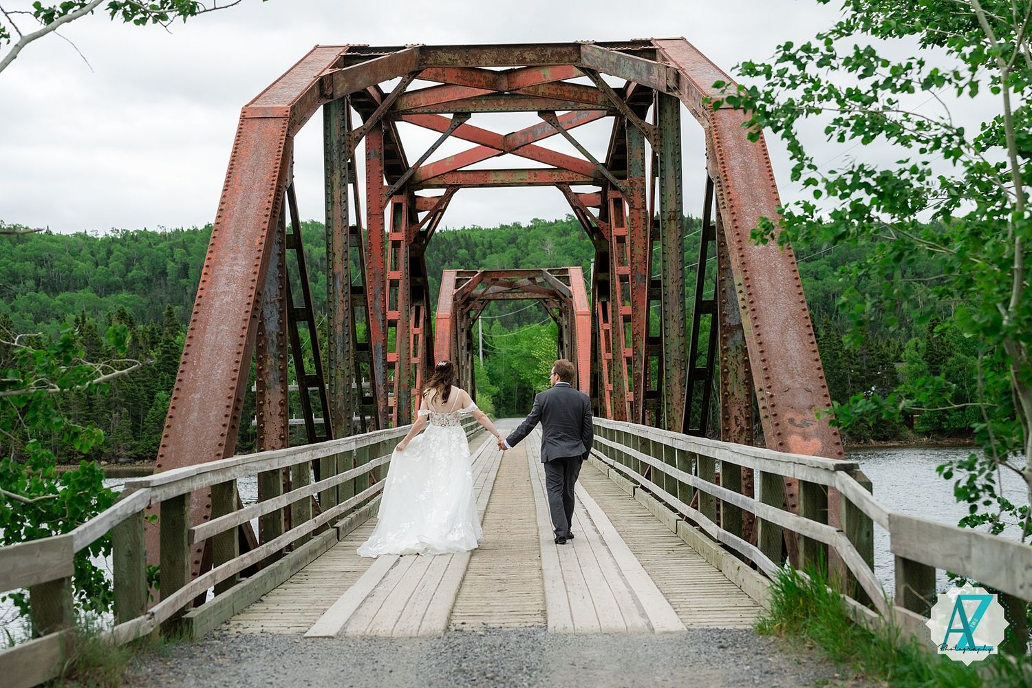 Two people walk across a rustic steel and wooden bridge surrounded by green trees and nature.