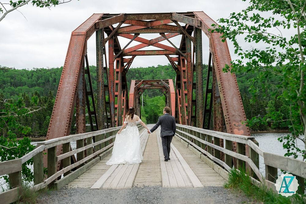 Two people walk across a rustic steel and wooden bridge surrounded by green trees and nature.