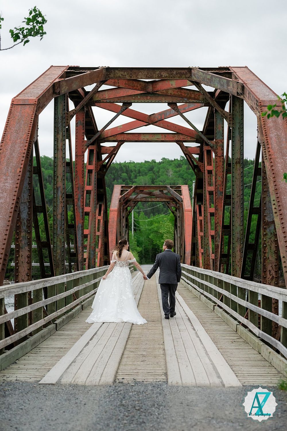 A couple walks along a wooden bridge with rustic red steel truss structure in a scenic outdoor setting.