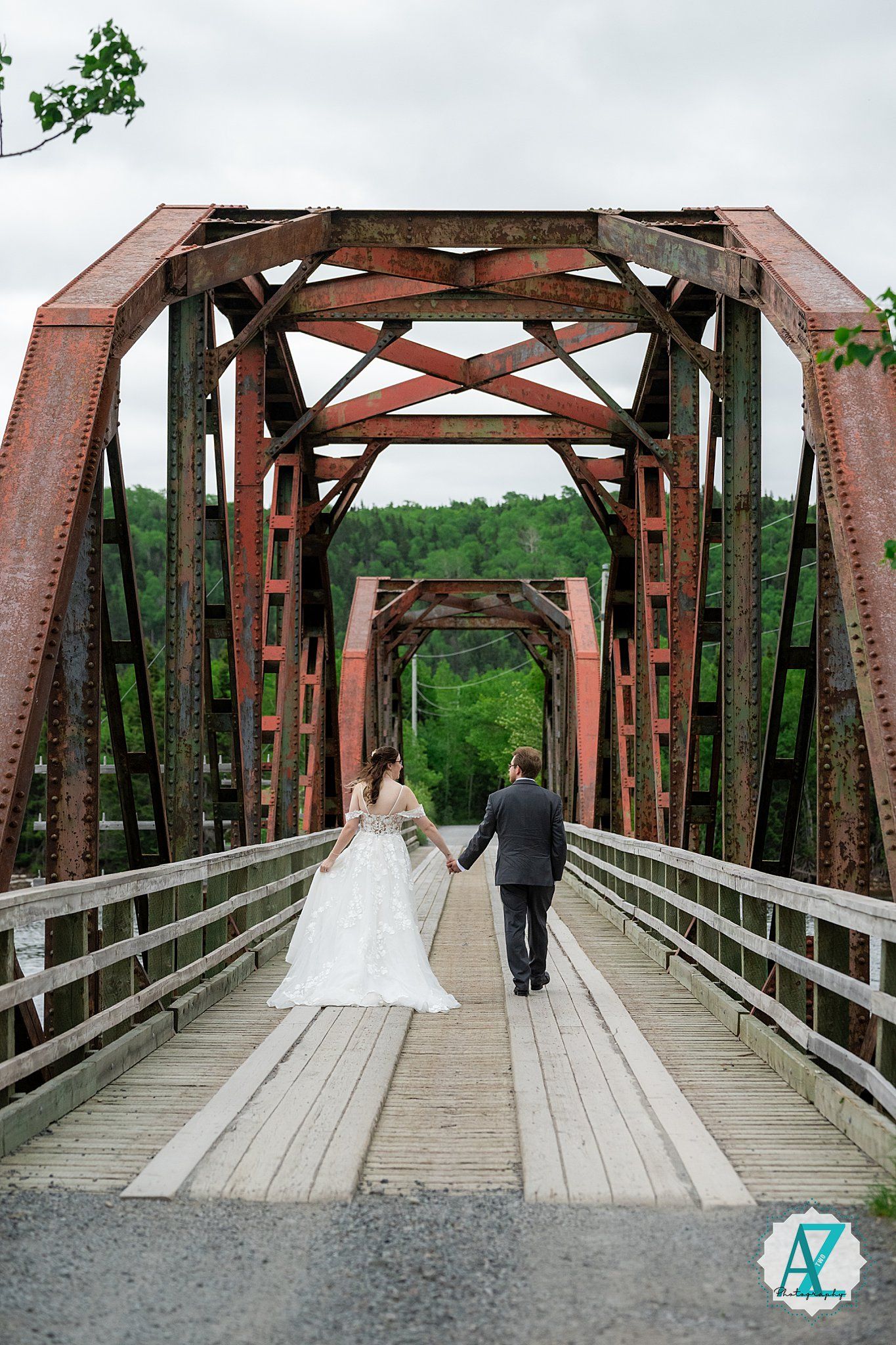 A couple walks along a wooden bridge with rustic red steel truss structure in a scenic outdoor setting.