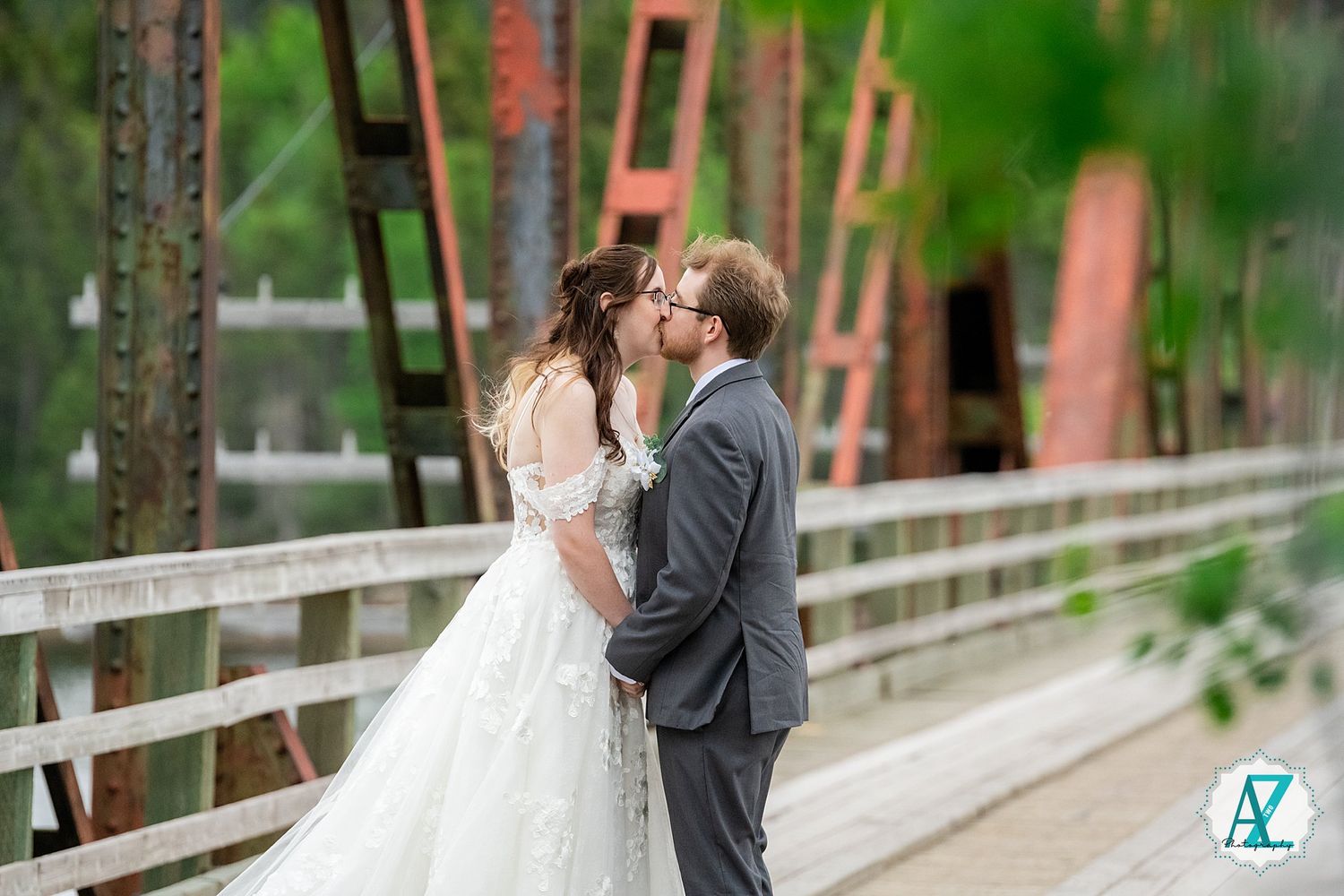 A couple in wedding attire share an intimate moment while standing on a wooden bridge surrounded by greenery.