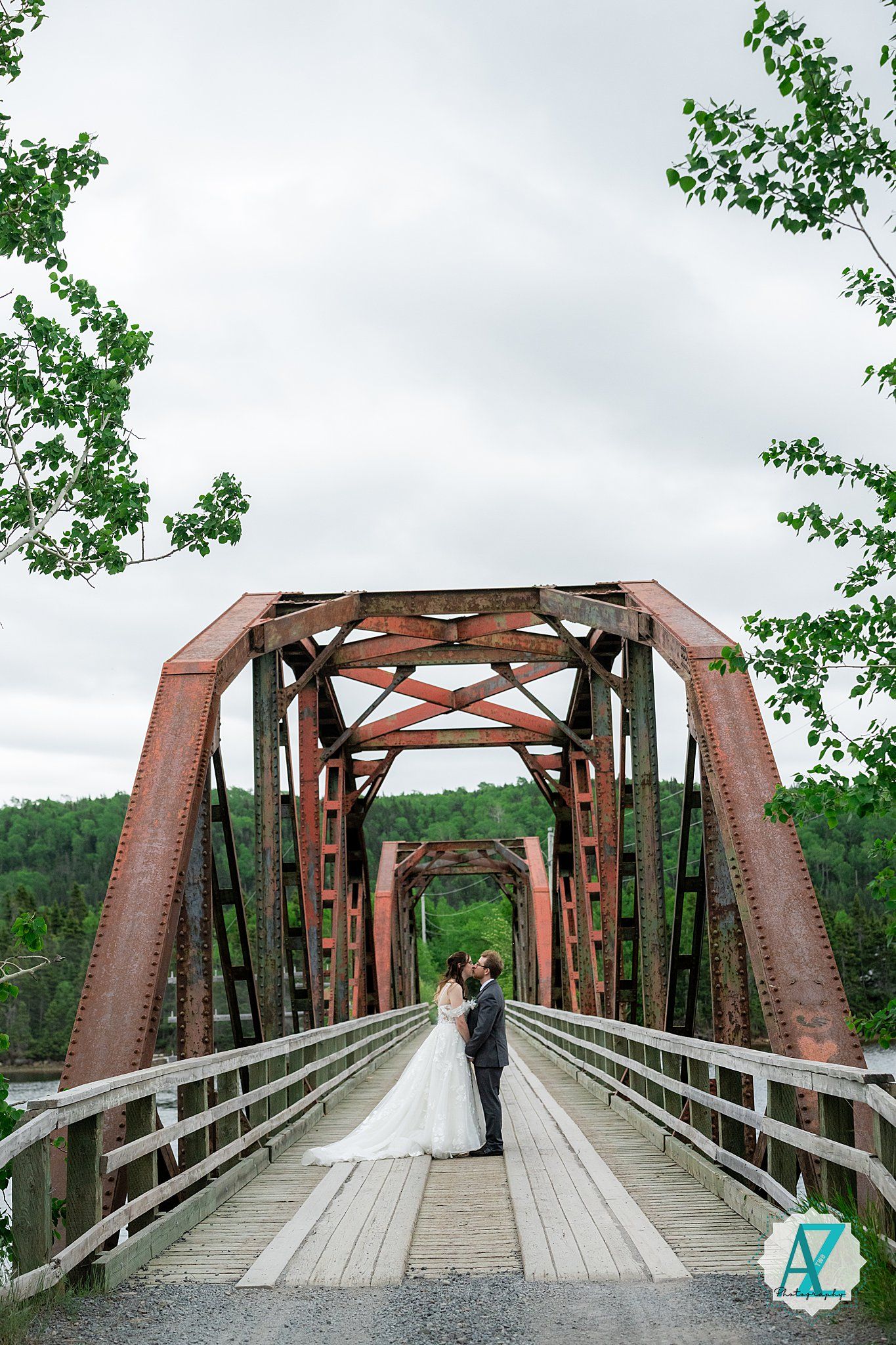 A rustic wooden bridge with red steel trusses spans over water in a scenic forest setting.