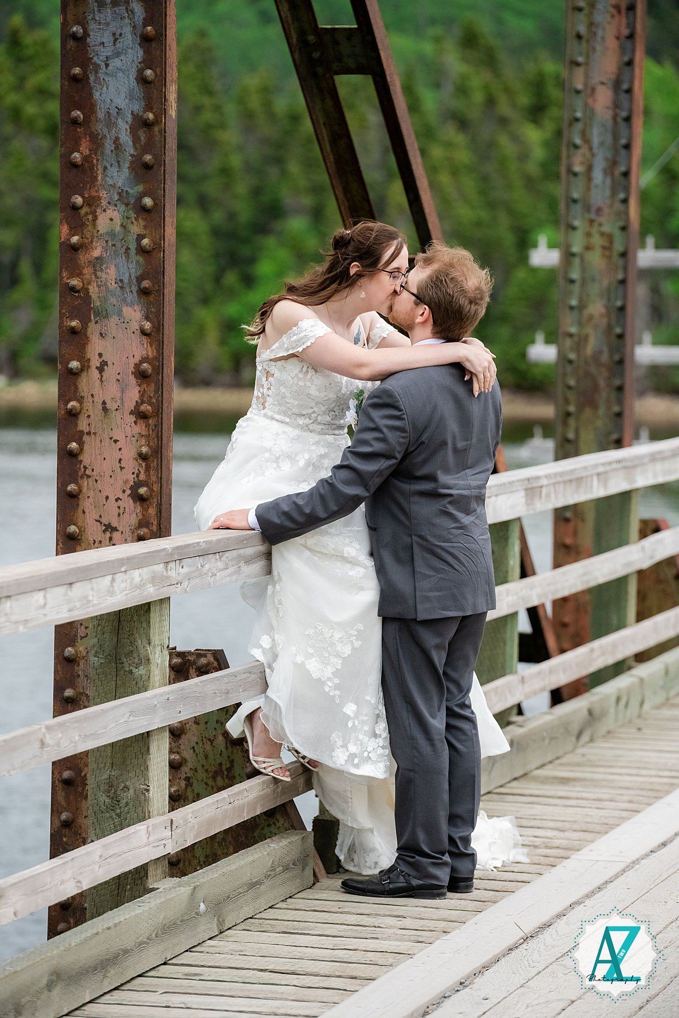 A romantic wedding couple embraces while sitting on a rustic wooden bridge over water.