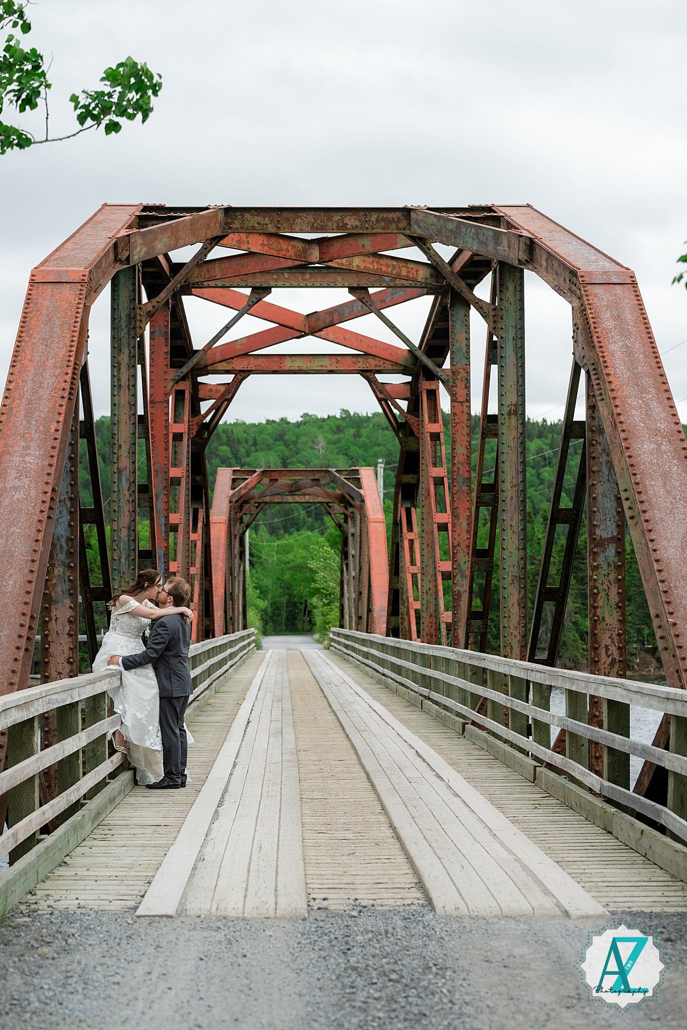 A rustic wooden boardwalk bridge with red metal trusses stretches across a scenic forested area on a cloudy day.