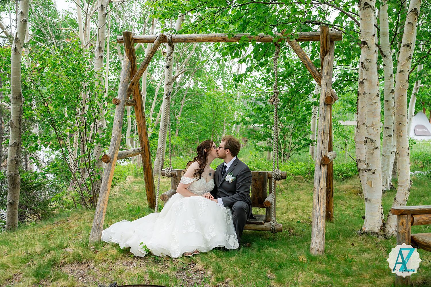 A couple shares a romantic moment on a rustic wooden swing under a birch tree arch in a woodland setting.