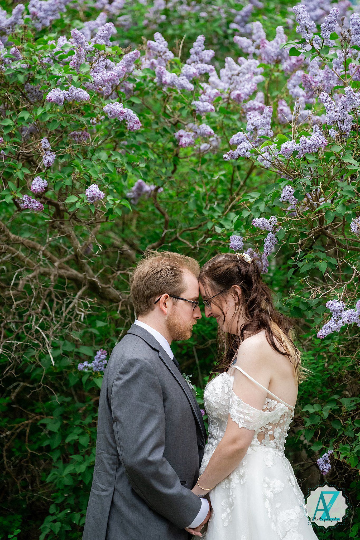 A couple shares an intimate moment beneath blooming purple lilac bushes in a garden setting.