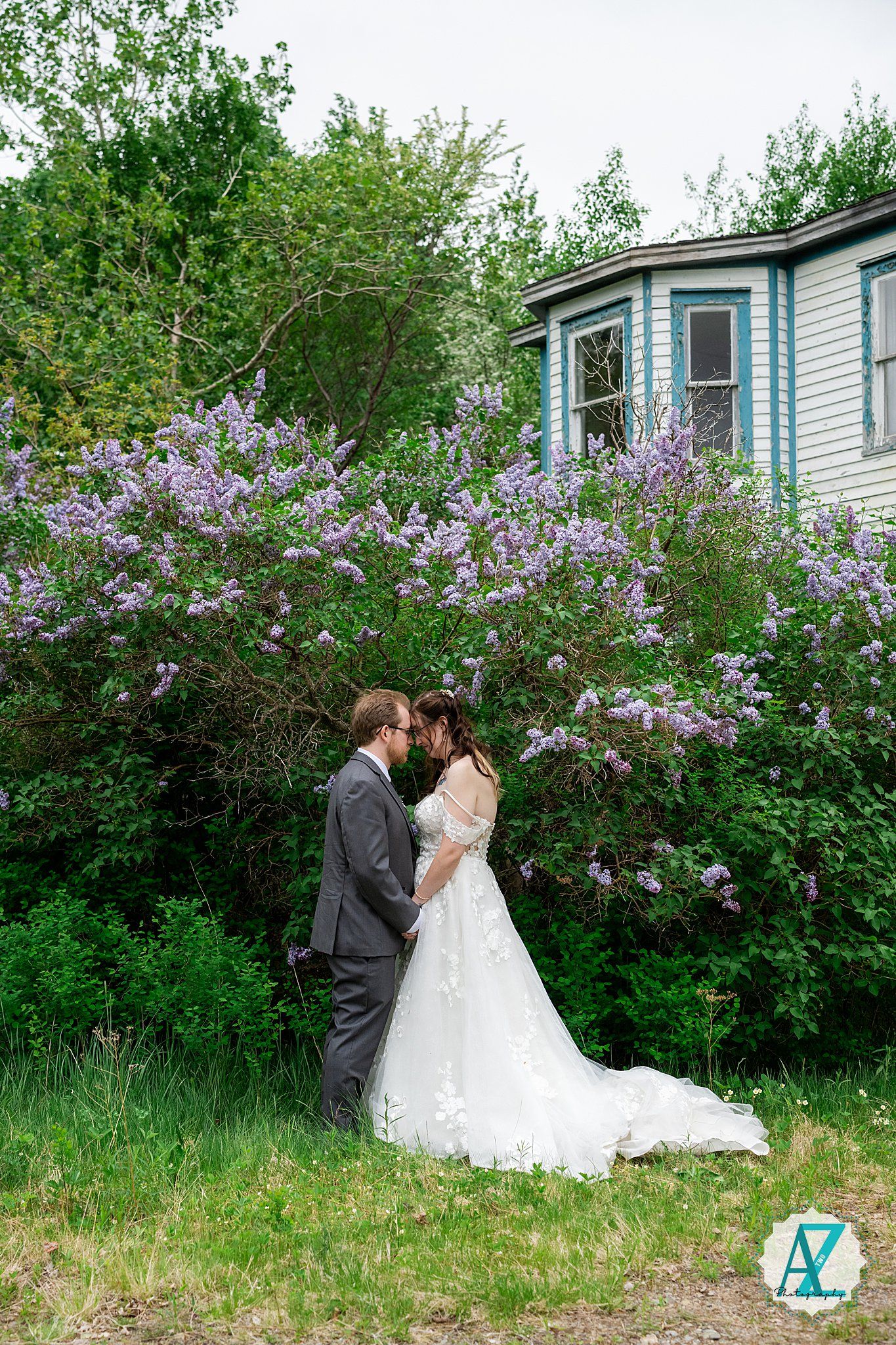 A couple shares a romantic moment in front of blooming lilac bushes by a light blue house on a spring day.