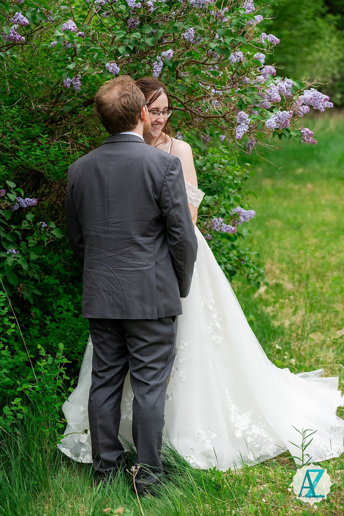 A couple in wedding attire stands together beside blooming lilac bushes in a garden setting.
