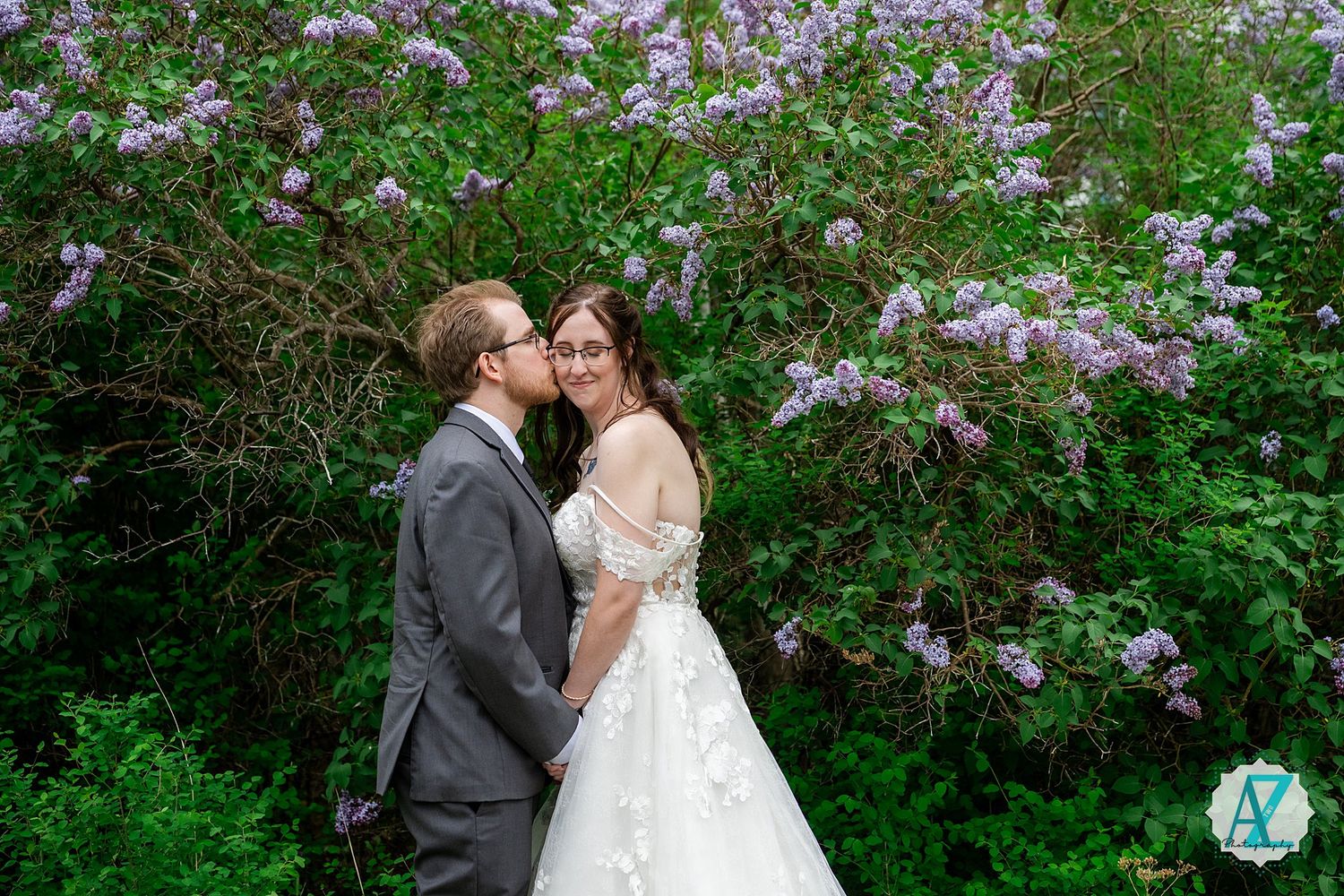 A couple shares a romantic kiss outdoors surrounded by blooming purple lilac bushes on a spring day.