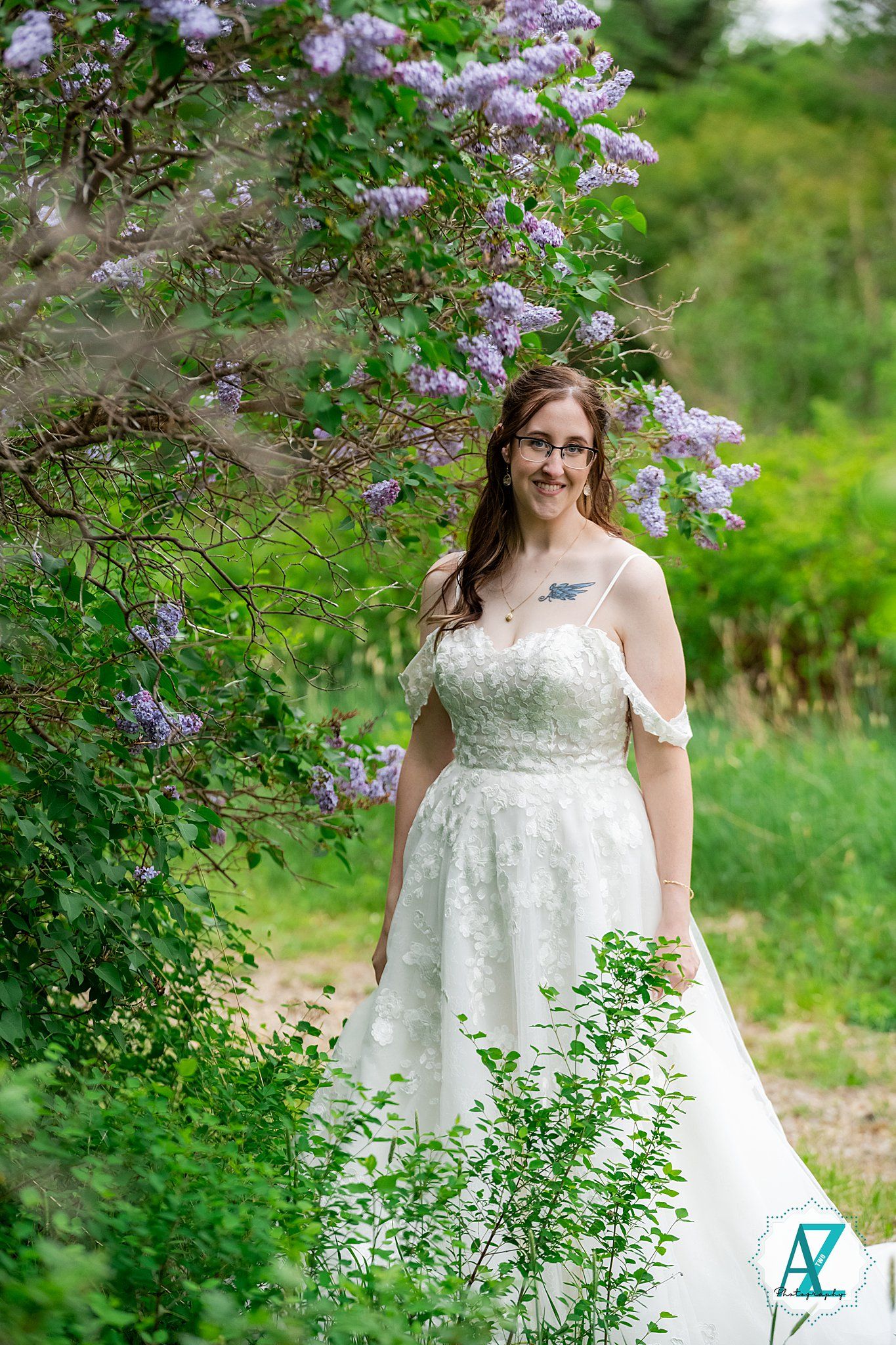 A white wedding dress with off-shoulder details stands beautifully framed by blooming purple lilac bushes in a lush garden setting.