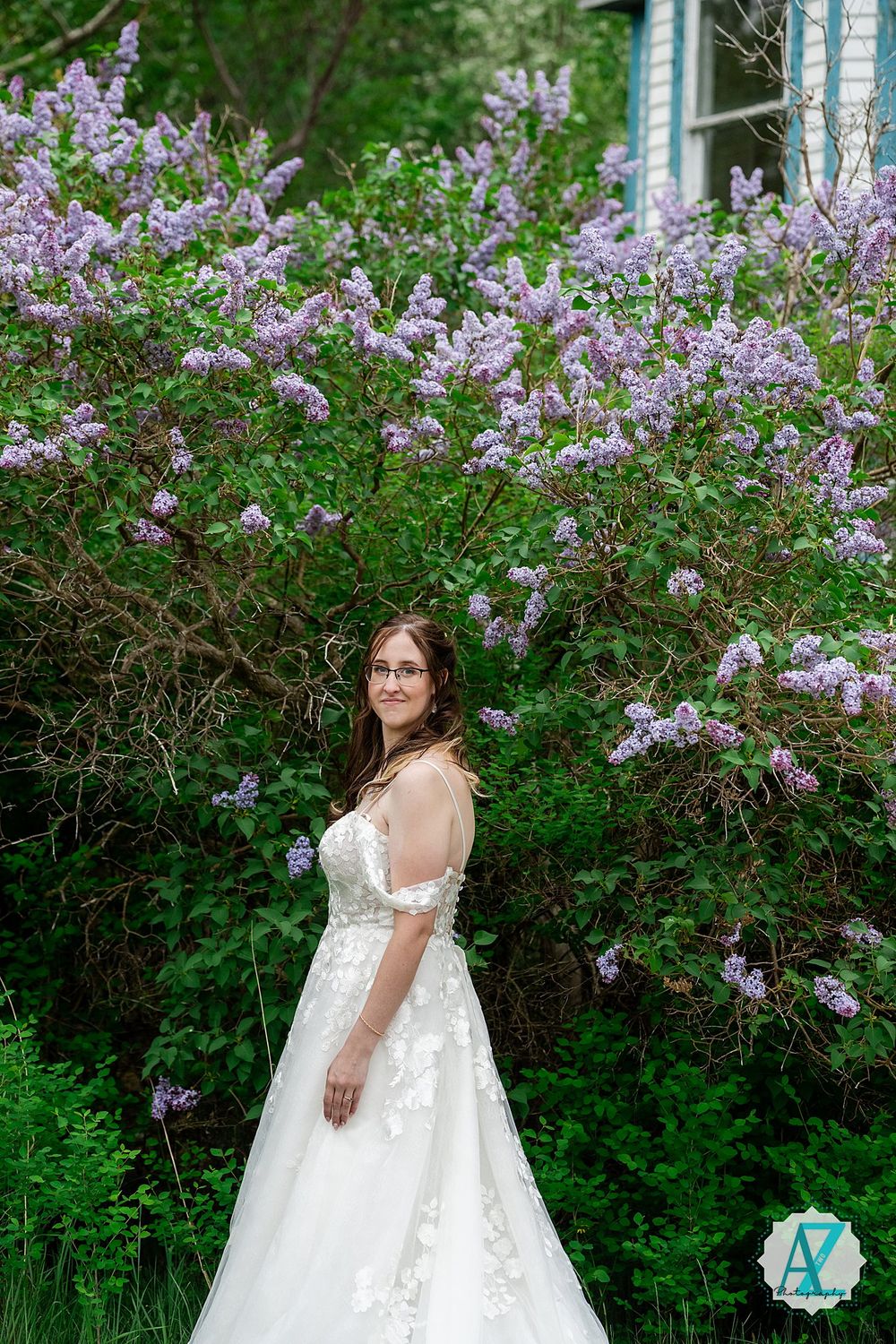 Elegant white wedding dress with floral lace details photographed against blooming purple lilac bushes in a garden setting.
