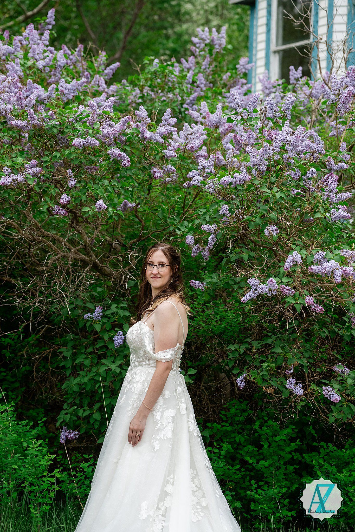 Elegant white wedding dress with floral lace details photographed against blooming purple lilac bushes in a garden setting.