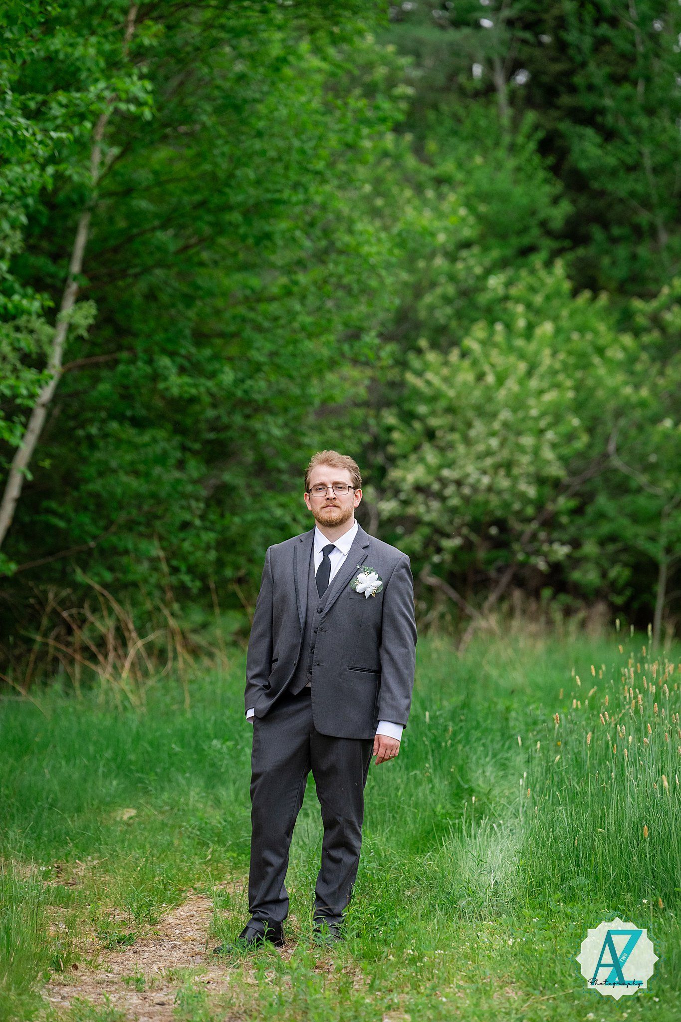 A groom in a charcoal gray suit stands on a dirt path surrounded by lush green forest foliage.