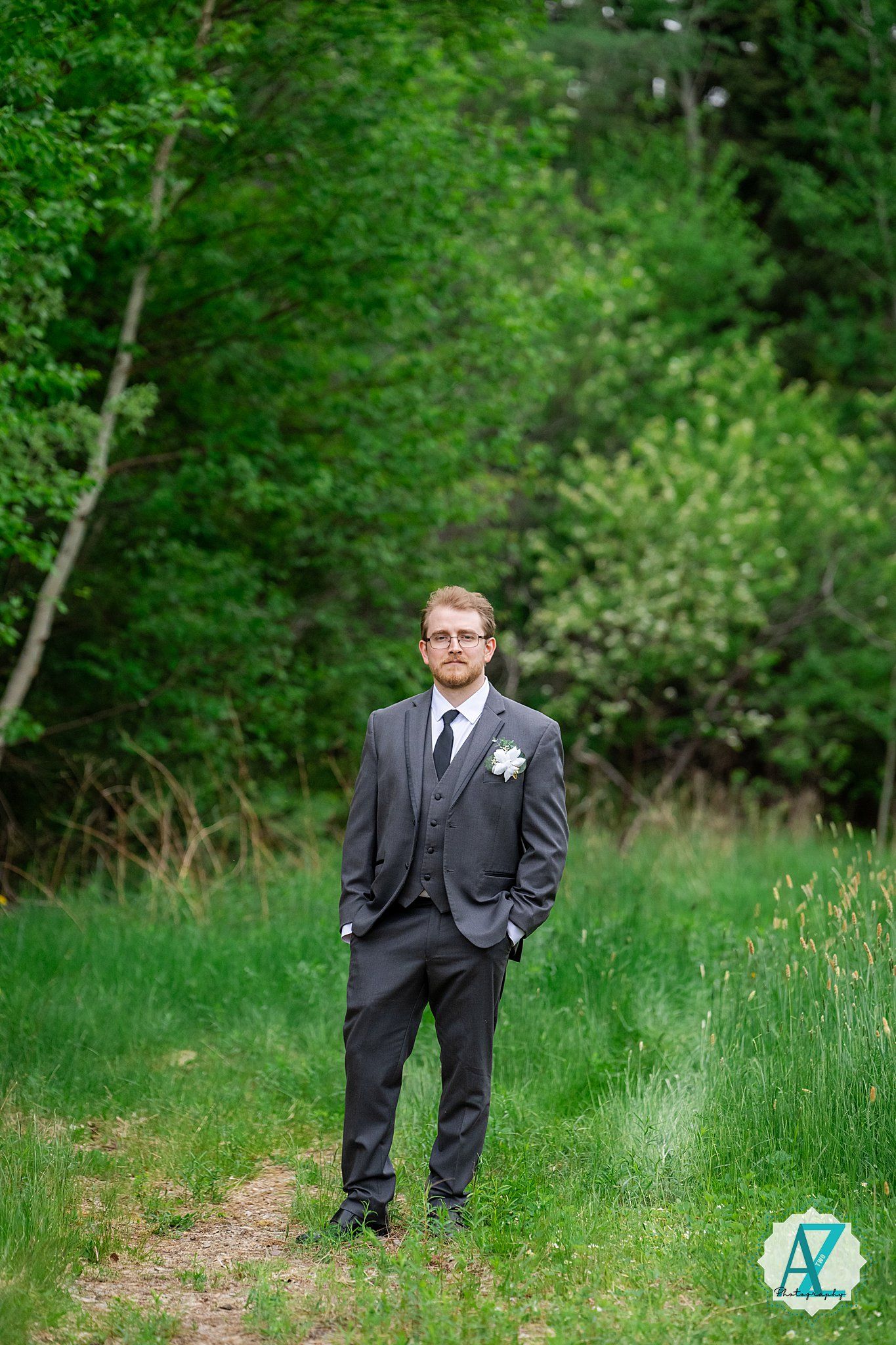 A groom in a dark grey three-piece suit poses confidently on a wooded trail surrounded by lush green foliage.