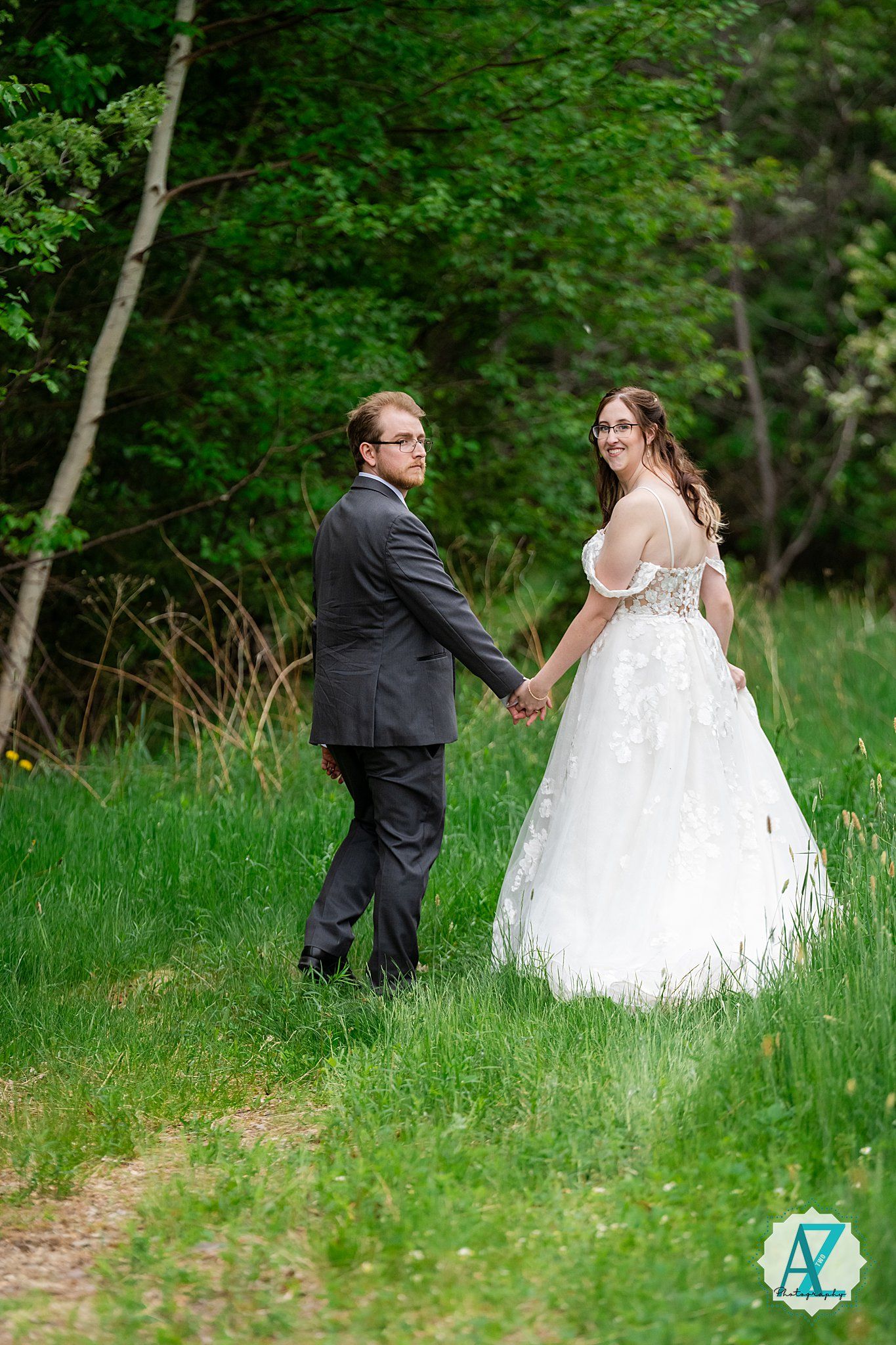 A romantic springtime wedding photo with a couple holding hands on a wooded path surrounded by lush green foliage.