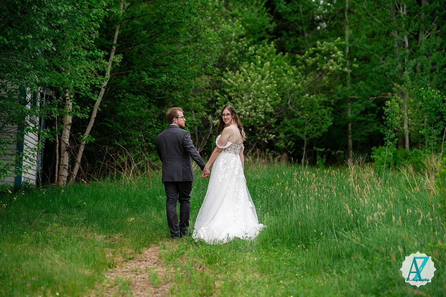 A couple in wedding attire walks together along a forest path surrounded by lush green trees and grass.