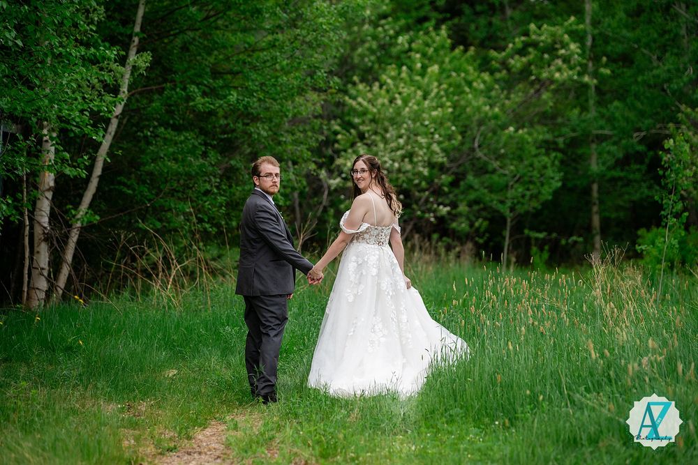 A couple stands together in a lush green meadow surrounded by trees on their wedding day.