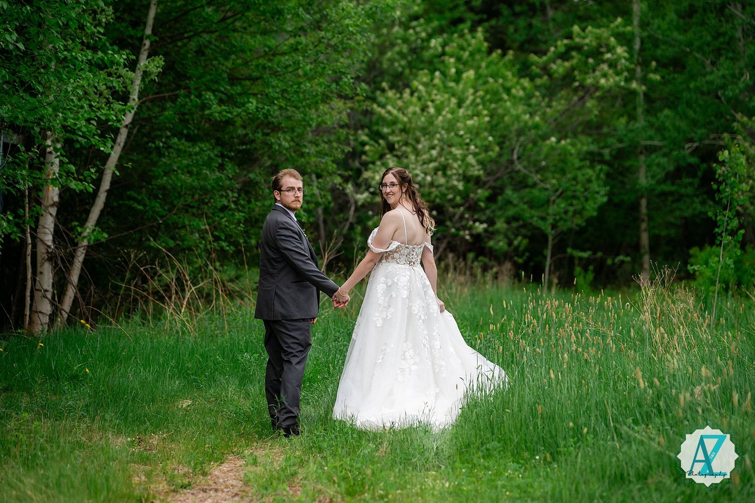A couple stands together in a lush green meadow surrounded by trees on their wedding day.