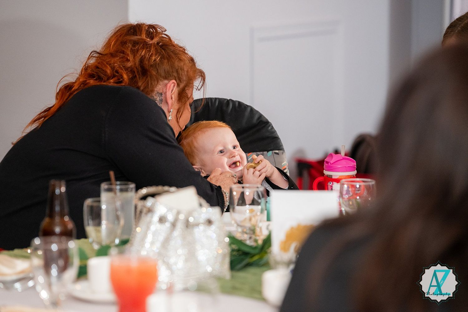 A baby being supported at a dining table with a festive meal setting and colorful drinks visible.