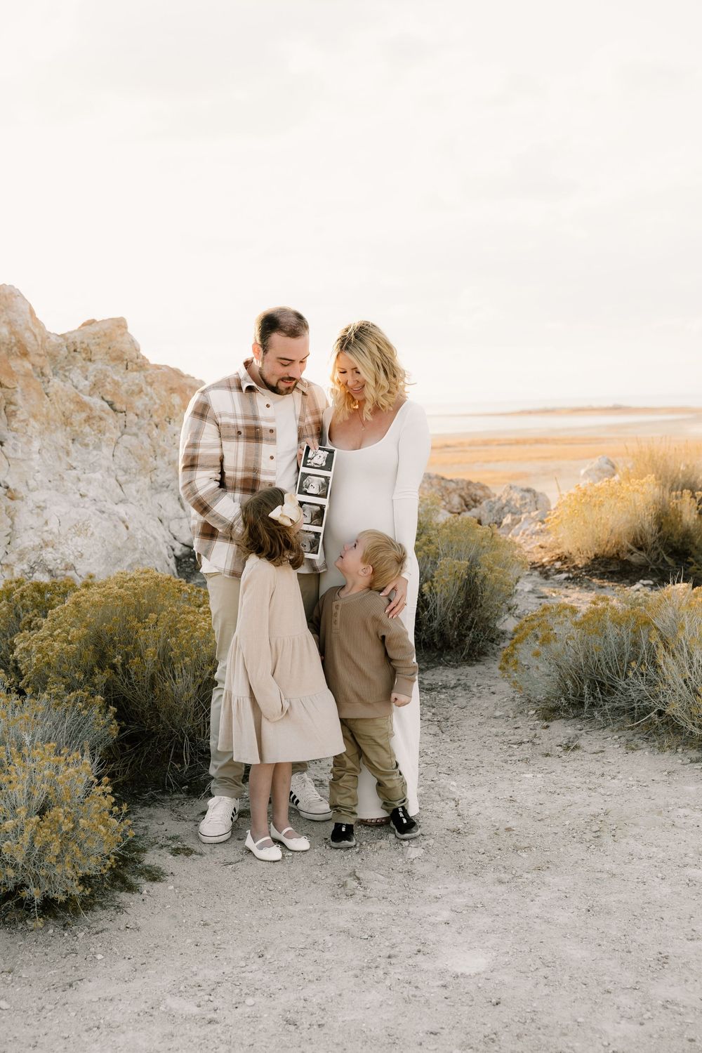 A casual family photoshoot in neutral tones set against a desert landscape at golden hour.