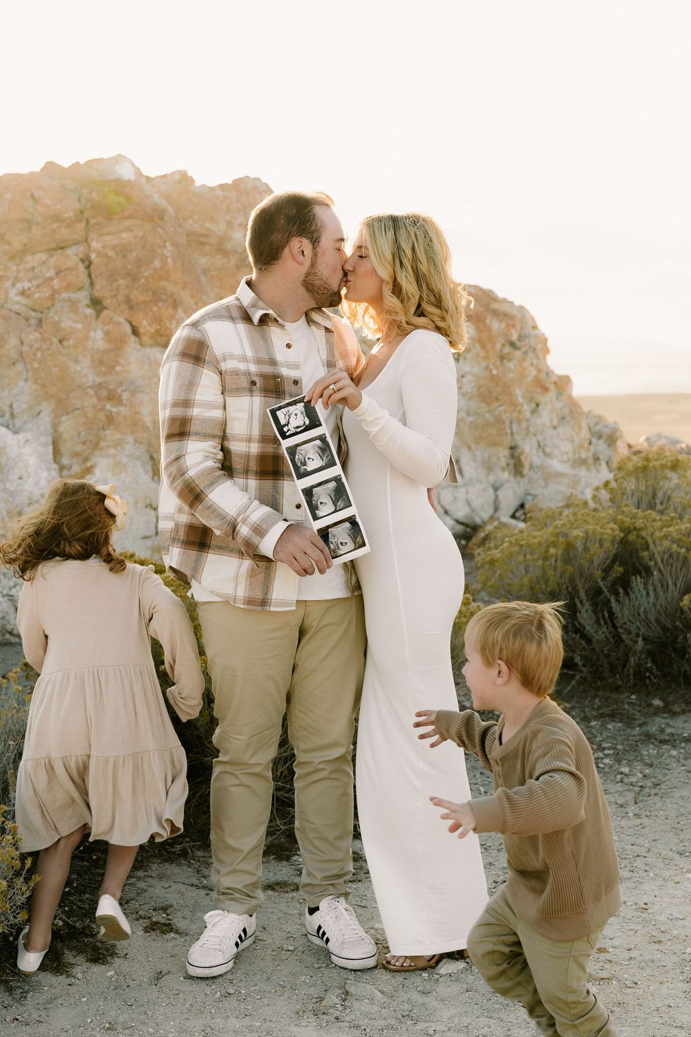 Family celebrating pregnancy announcement with ultrasound photos on beach during golden hour sunset session.