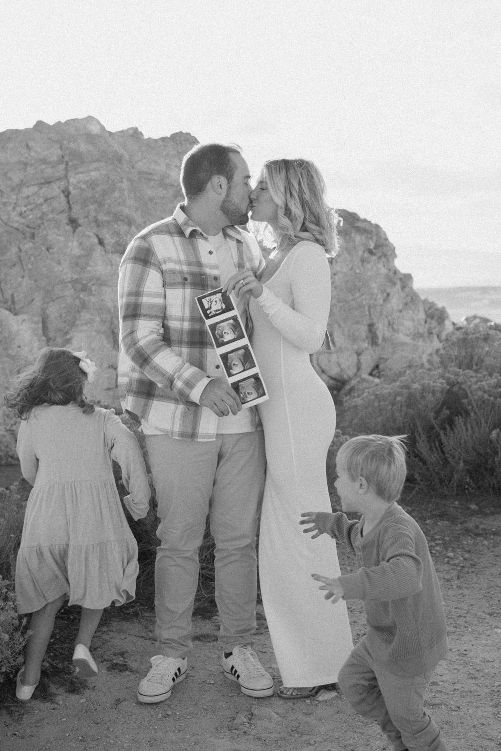 Black and white family photo in a rocky desert setting showing a pregnancy announcement with sonogram pictures.