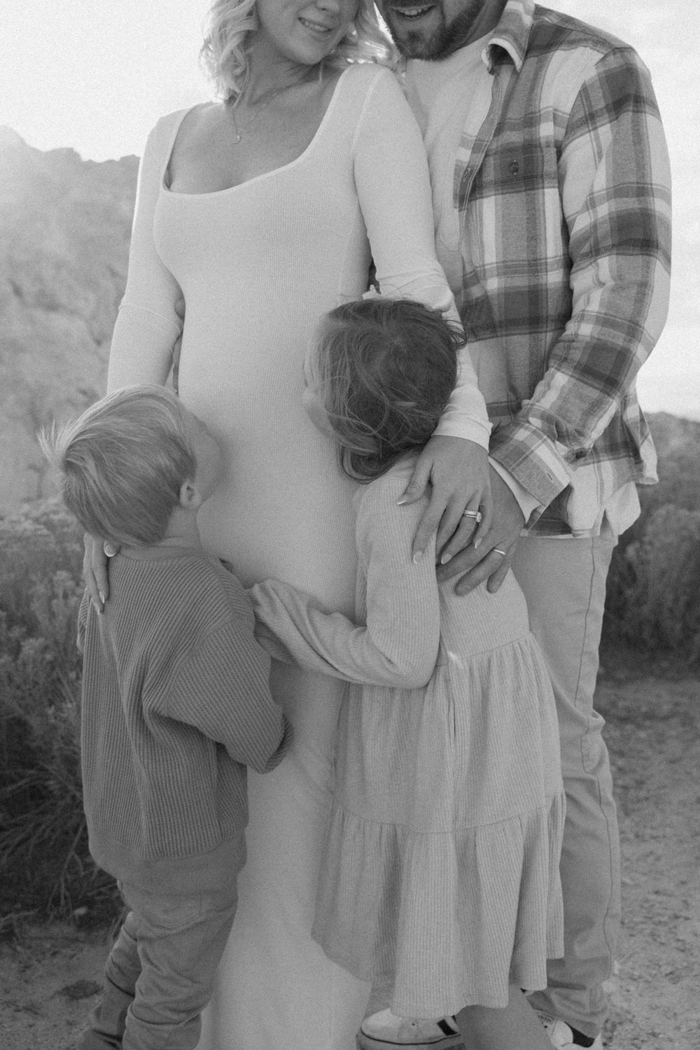 A tender black and white family photo showing a pregnant woman in a white dress with loved ones embracing outdoors.