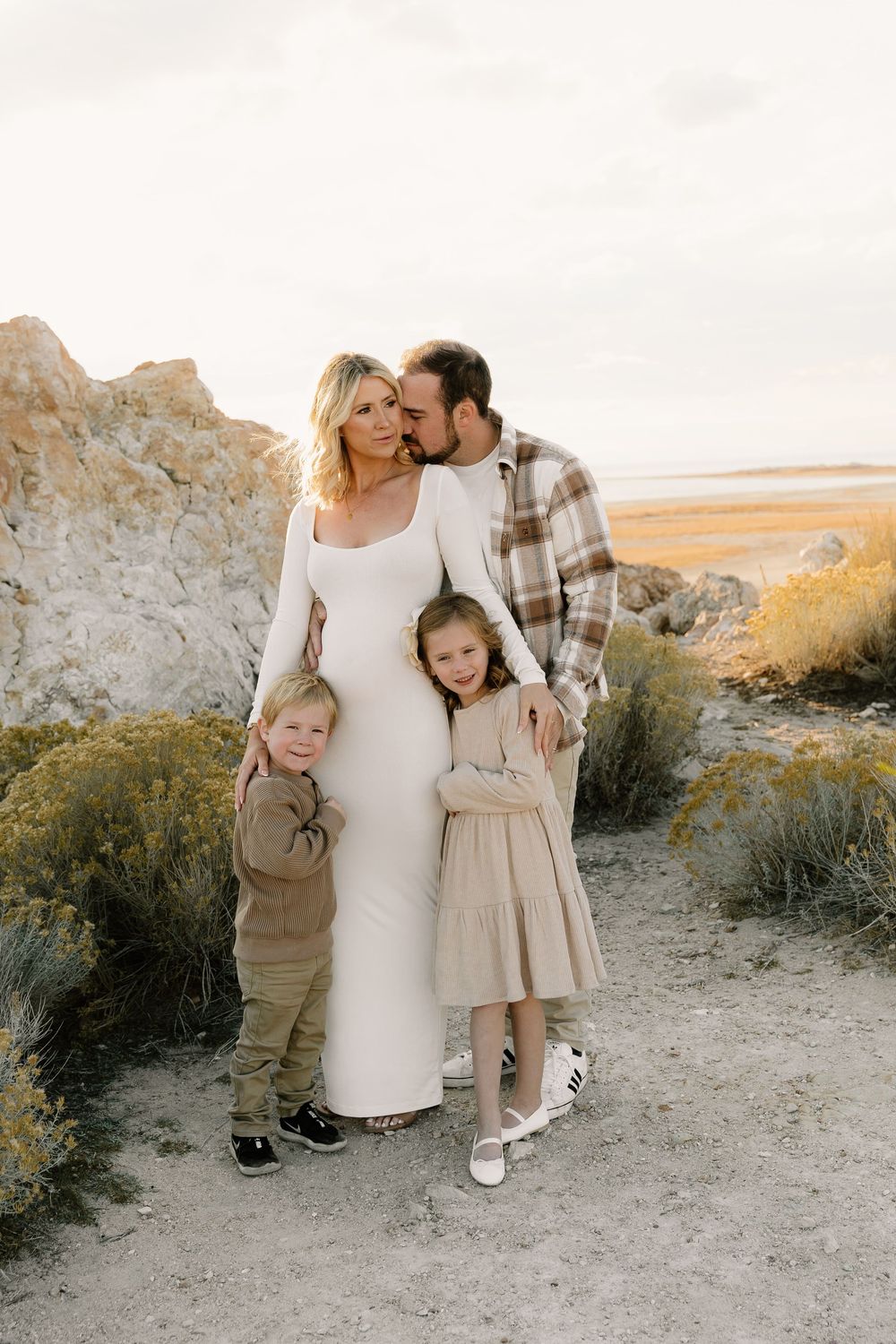 Family poses together in desert landscape wearing neutral tones against rocky formations at sunset.