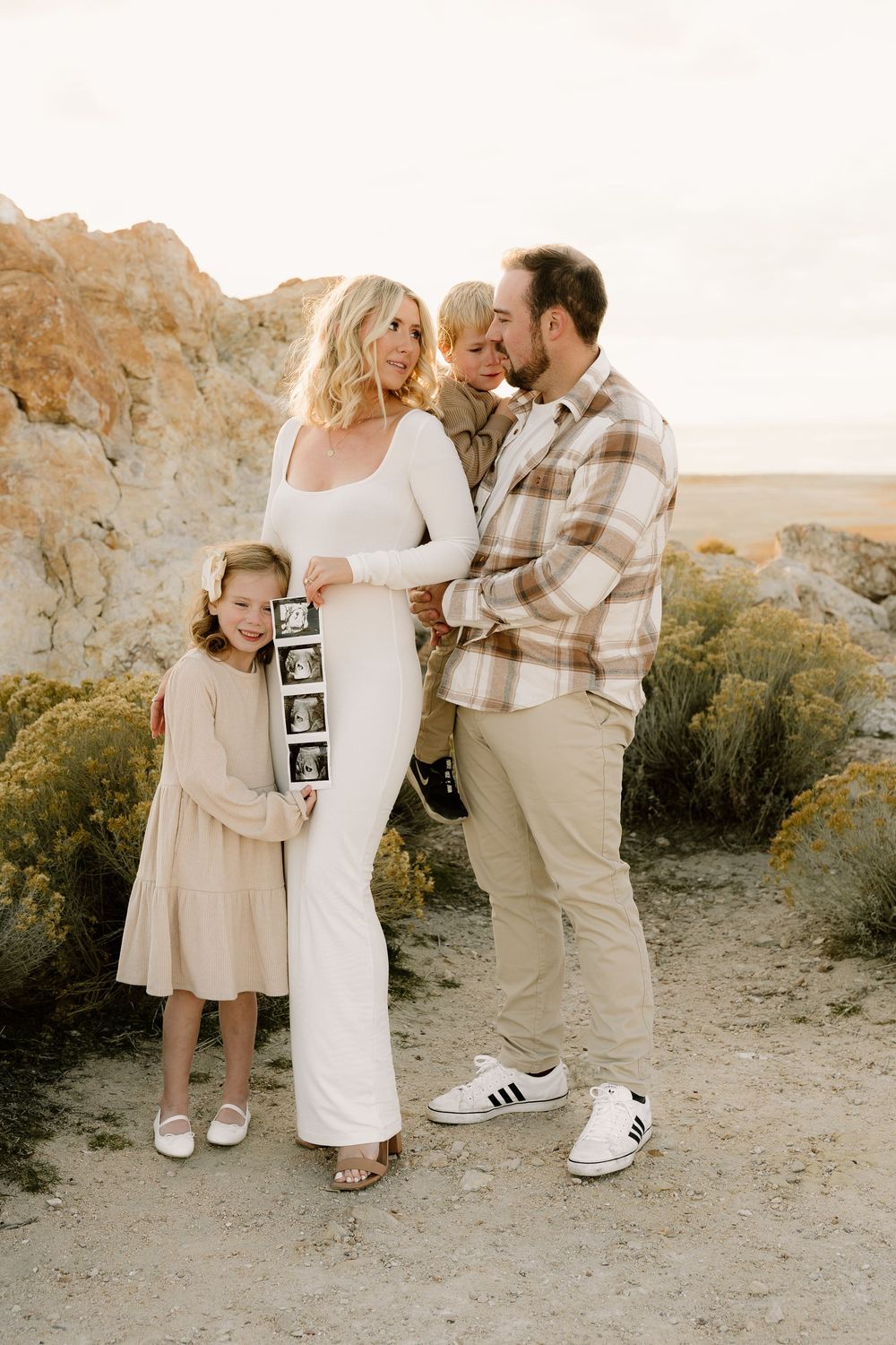 A family pregnancy announcement photo captured in a desert landscape at sunset with ultrasound images.