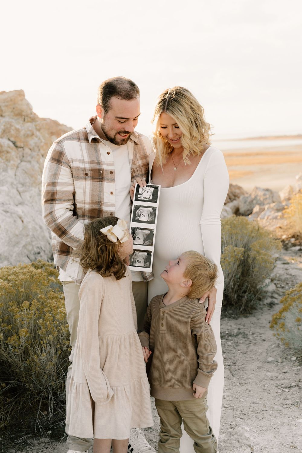 A family celebrates their pregnancy announcement in a desert landscape while holding ultrasound photos.