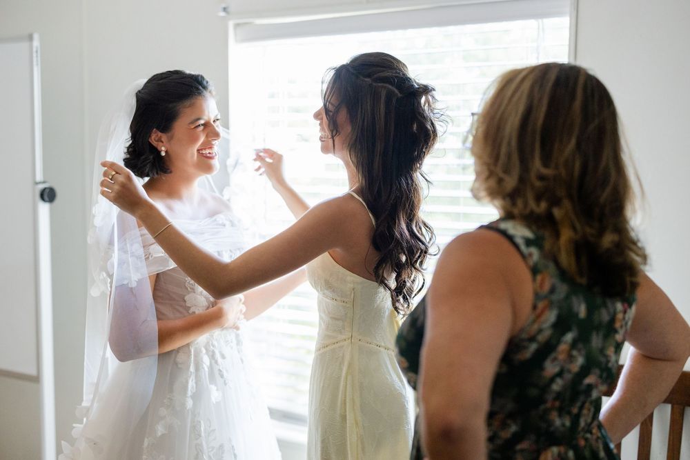 Bride getting help with her wedding dress during preparation with bridesmaids in bright room.