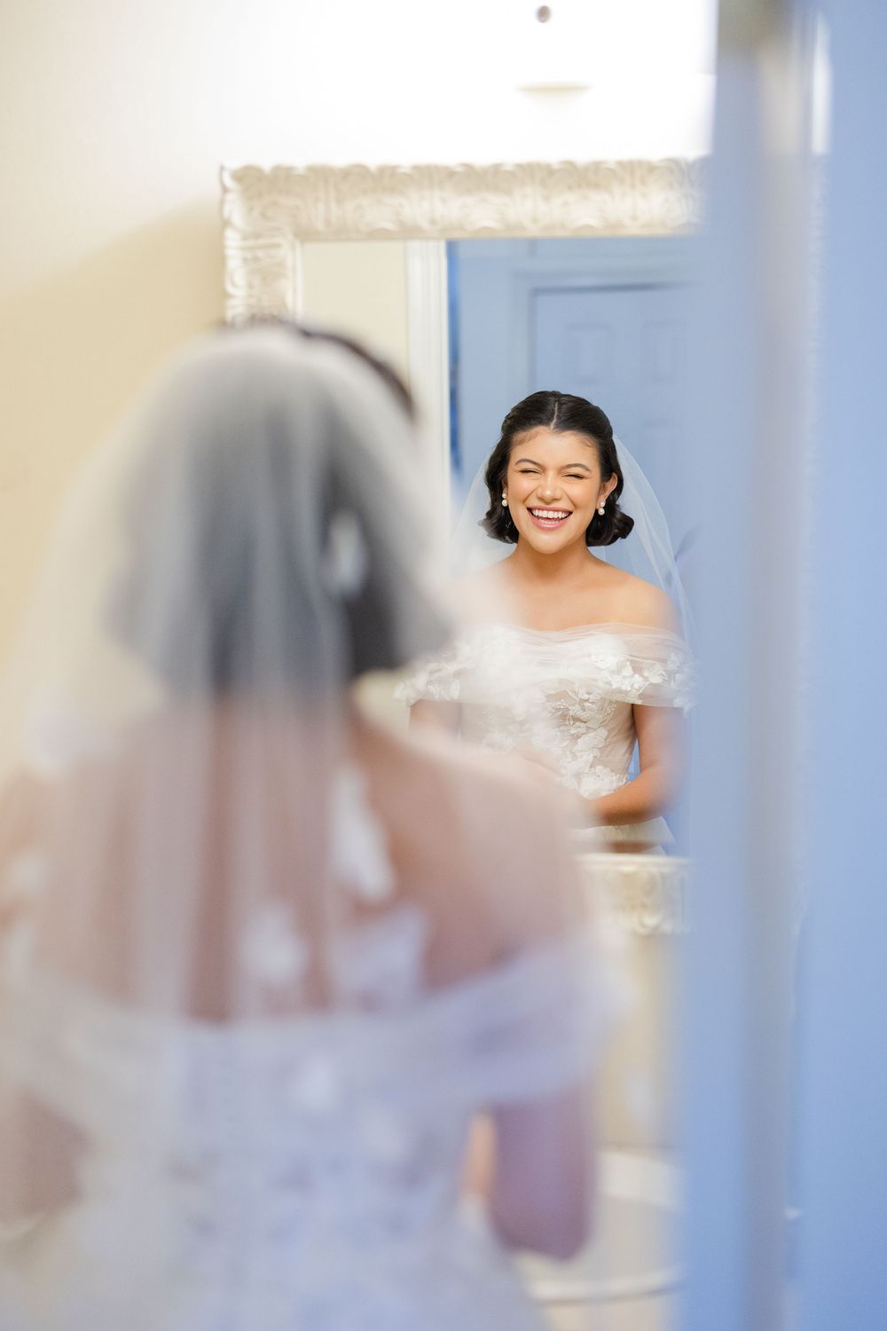 A bride wearing an off-shoulder white wedding dress smiles joyfully while looking at her reflection in a mirror.