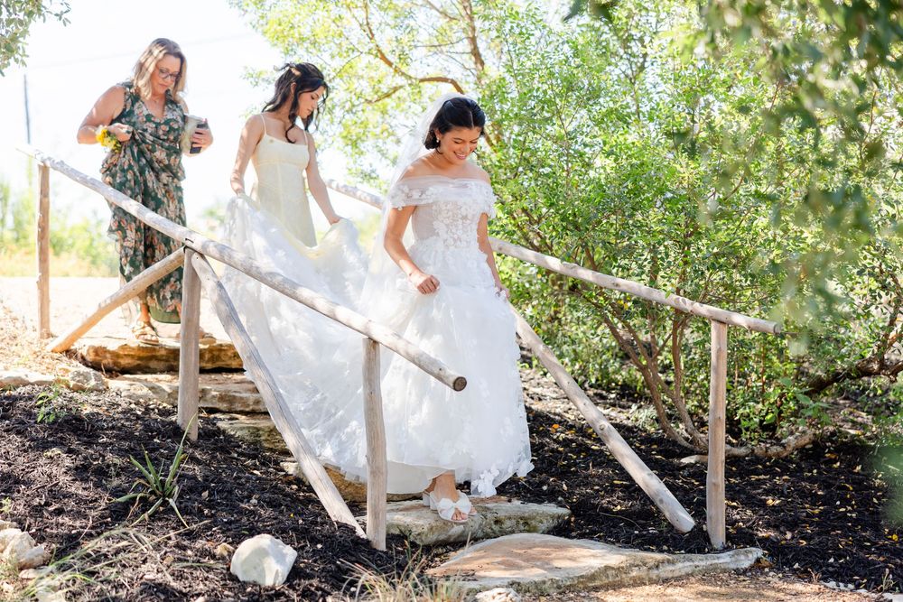 Three people in white dresses carefully walking down rustic wooden steps surrounded by greenery.