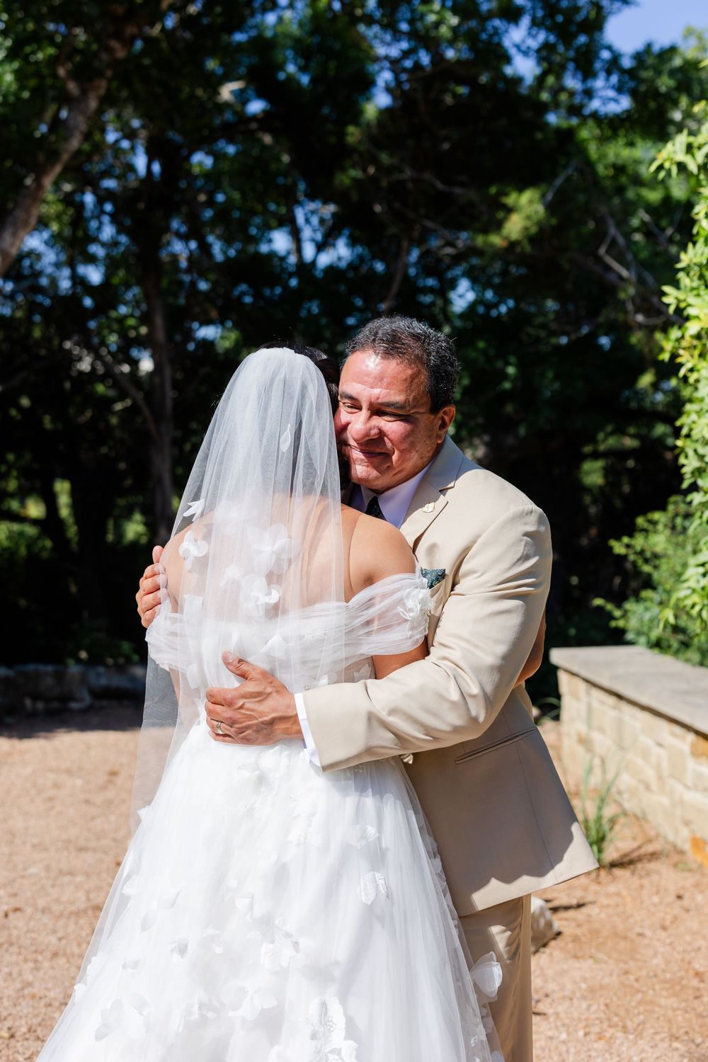 A heartfelt embrace between a bride in white and a man in tan suit during an outdoor wedding ceremony.