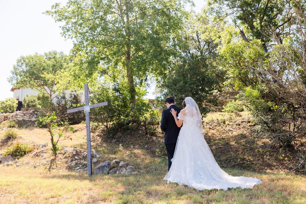 A wedding couple stands together on a sunny hillside with trees and greenery in the background.
